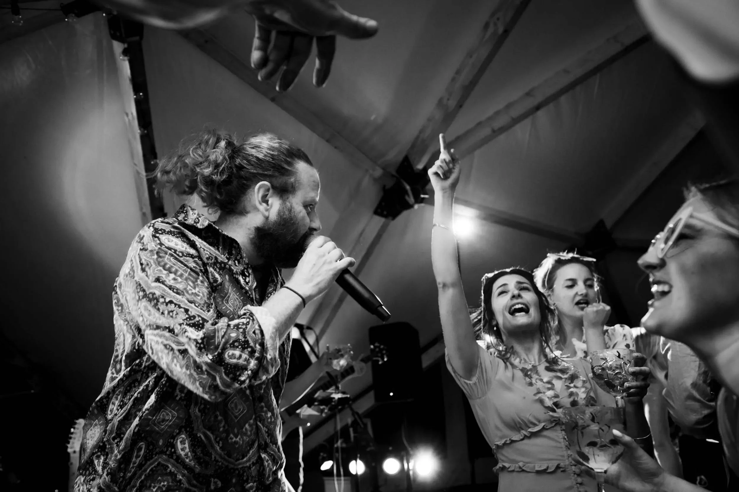 A man with long hair tied back singing into a microphone, surrounded by a group of women at a lively party or concert in a tent, one woman with sunglasses and a flower lei raising her hand, others holding drinks and enjoying the moment.