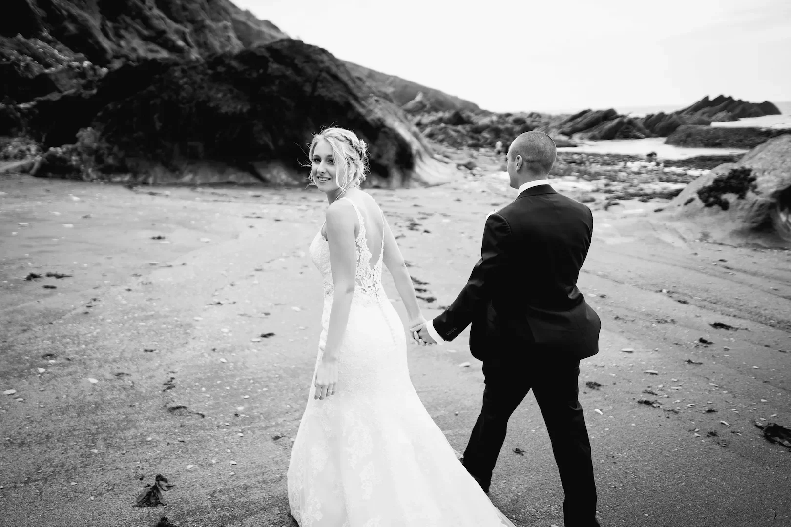 A black and white photo of a bride and groom walking hand in hand on a beach, with rocky formations in the background.