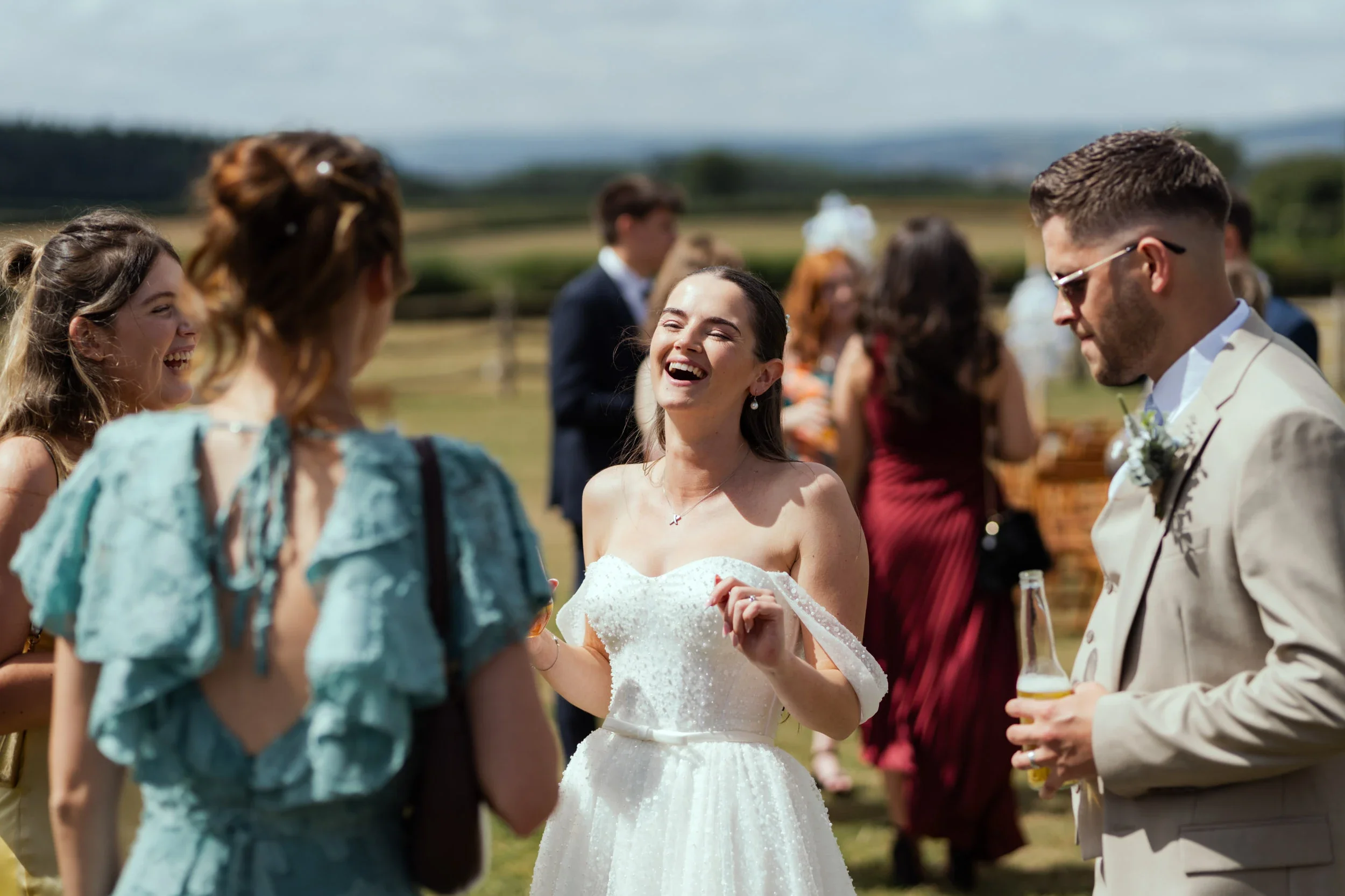 A group of people at an outdoor wedding celebration. The bride, wearing a white strapless dress, is smiling and laughing while talking to guests. The groom, in a light-colored suit, is holding a beverage and wearing sunglasses. Others in the backgrou