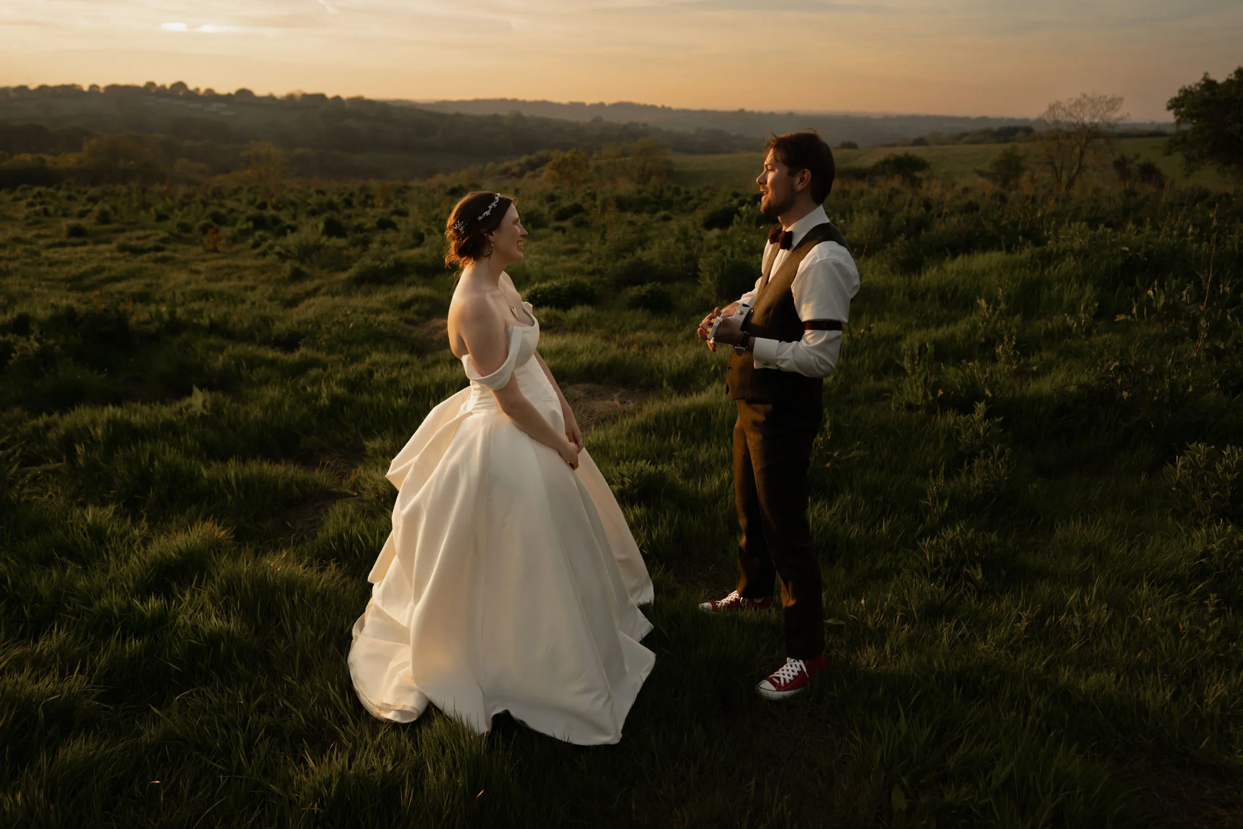 A bride and groom stand in a grassy field at sunset, facing each other. The bride wears a white wedding gown with off-the-shoulder sleeves, and the groom wears a dark vest and pants with sneakers. The scene has a warm, romantic ambiance with a scenic