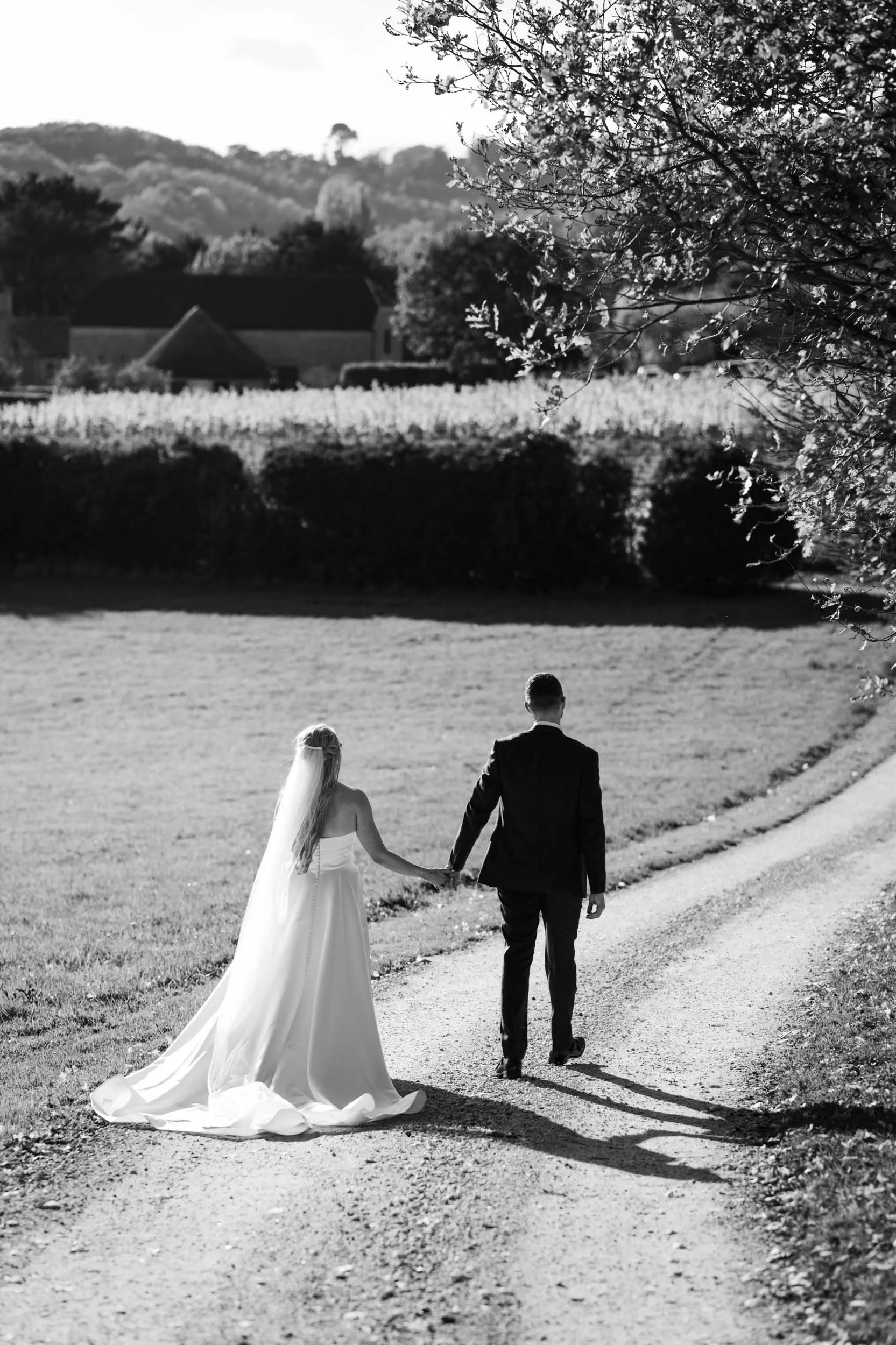 A black and white photo of a bride and groom holding hands and walking away on a dirt path in a rural setting.