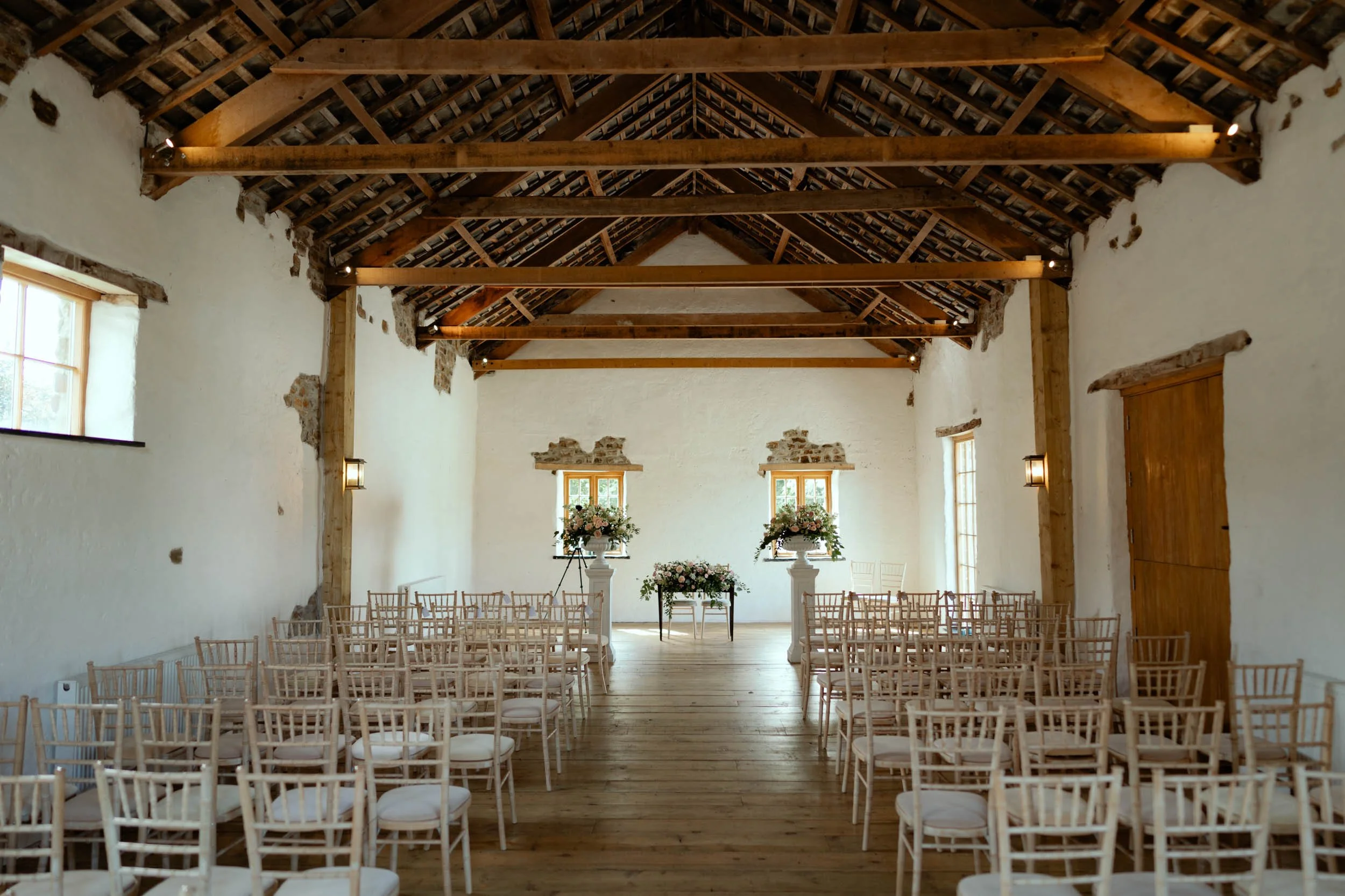 Empty wedding ceremony space with white chairs, floral arrangements, and a wooden ceiling in a rustic barn.