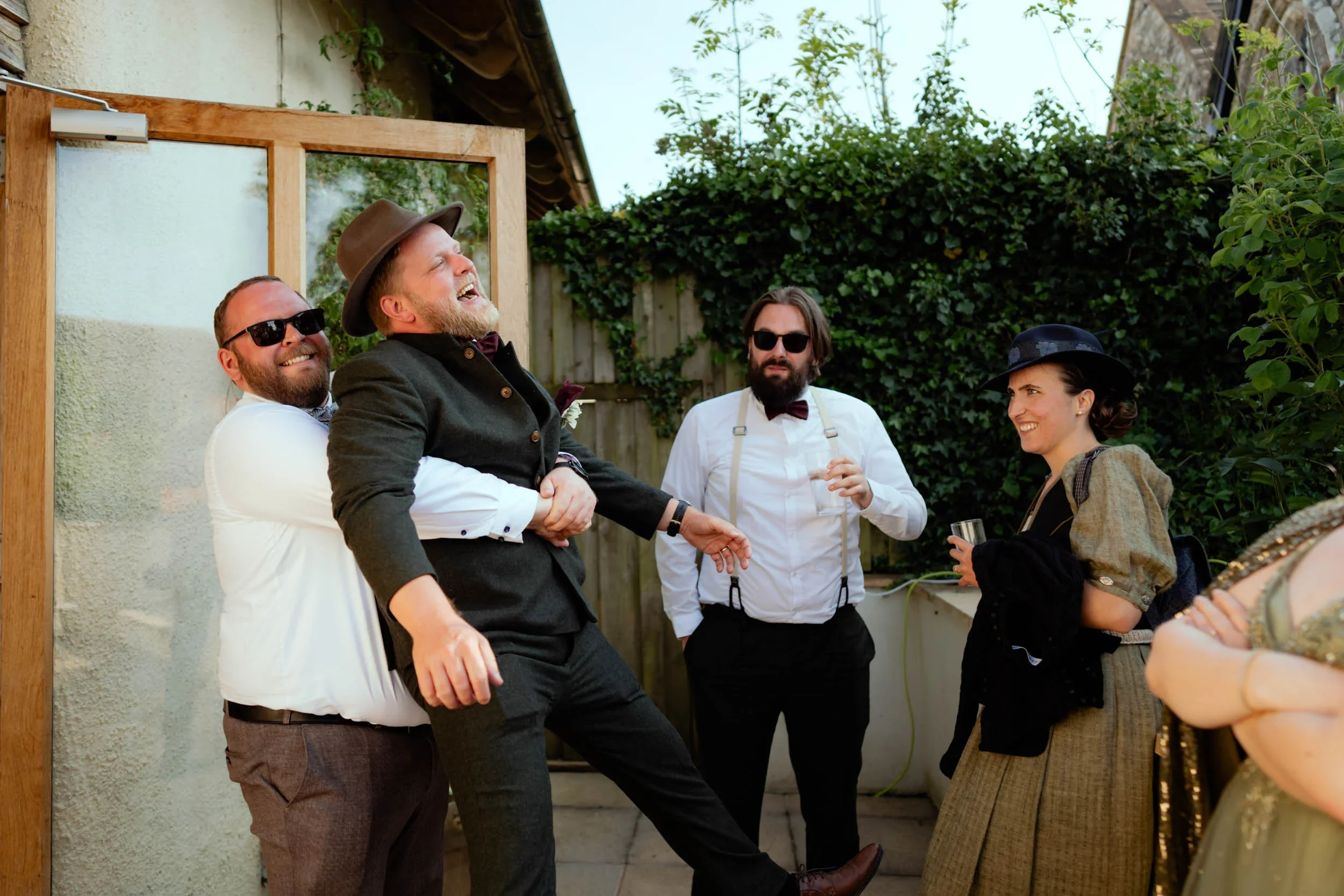 Group of five people at an outdoor gathering, one man being playfully held by another, all dressed in vintage or semi-formal clothing, wearing sunglasses, and smiling or talking with each other, with greenery and a fence in the background.