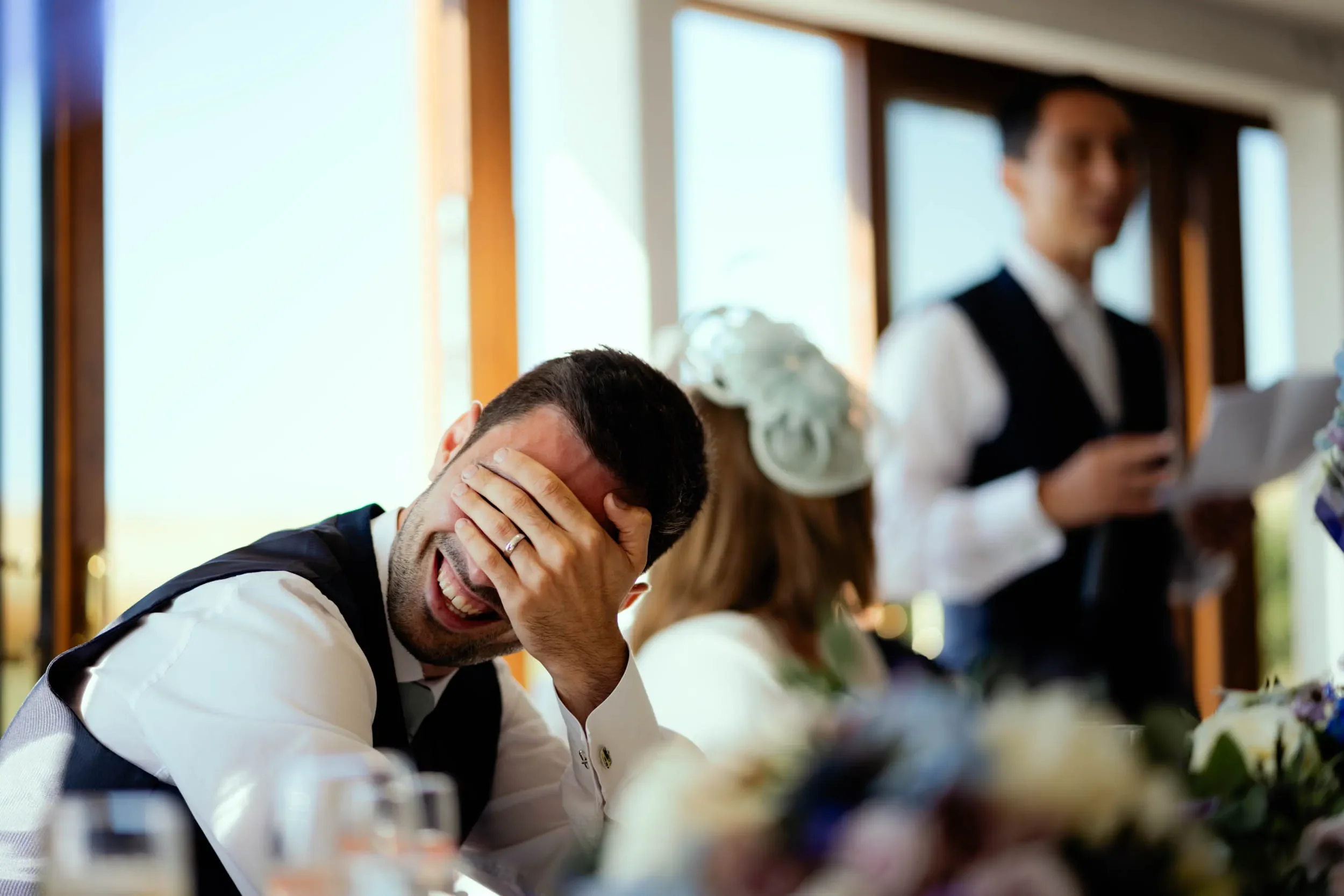 A man in formal attire laughing with his hand over his face at a celebration, seated at a table with flowers, with a woman wearing a white dress and floral hairpiece and a man in a vest and white shirt in the background.