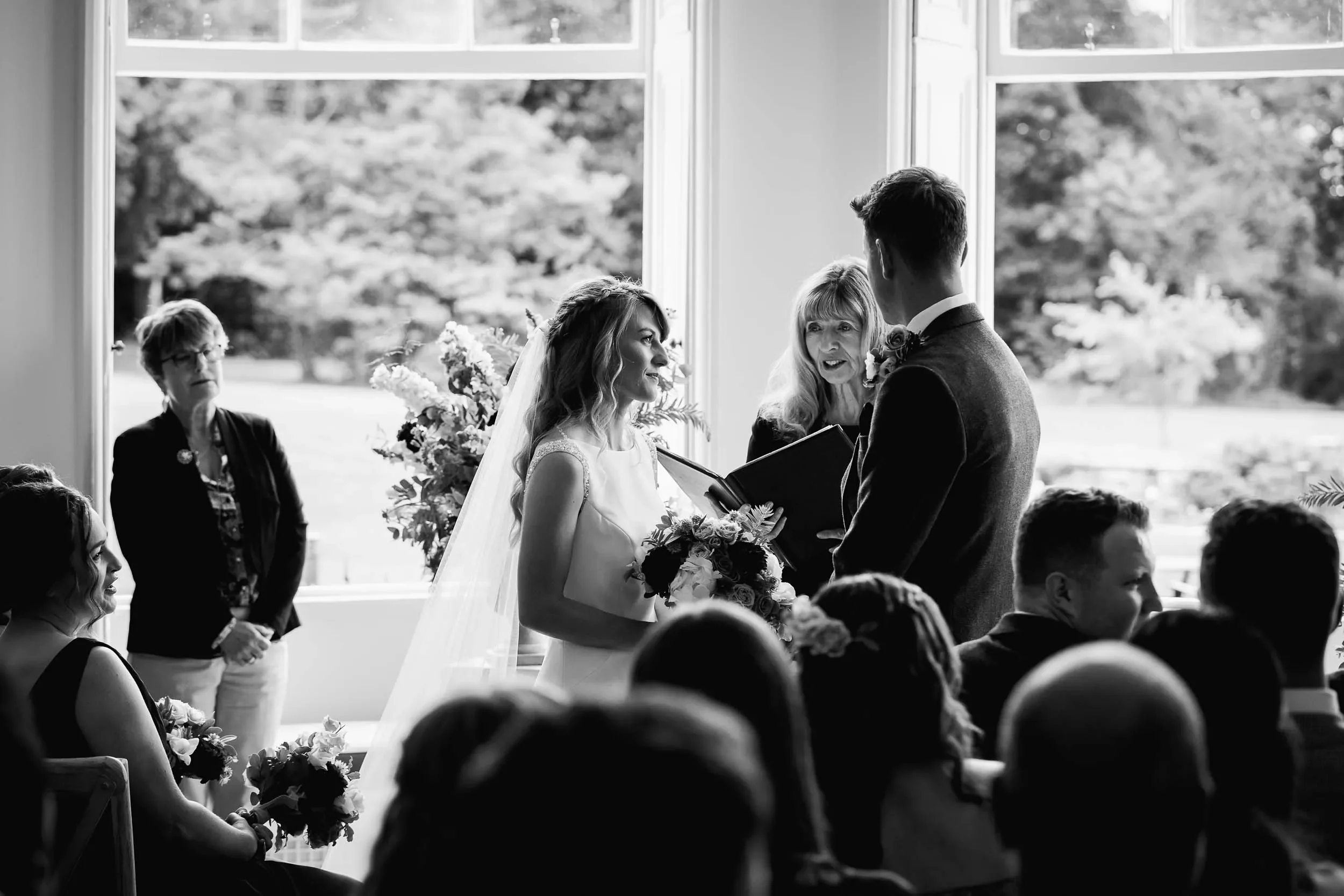 Black and white photo of a wedding ceremony with the bride and groom exchanging vows, officiant speaking, and guests watching.