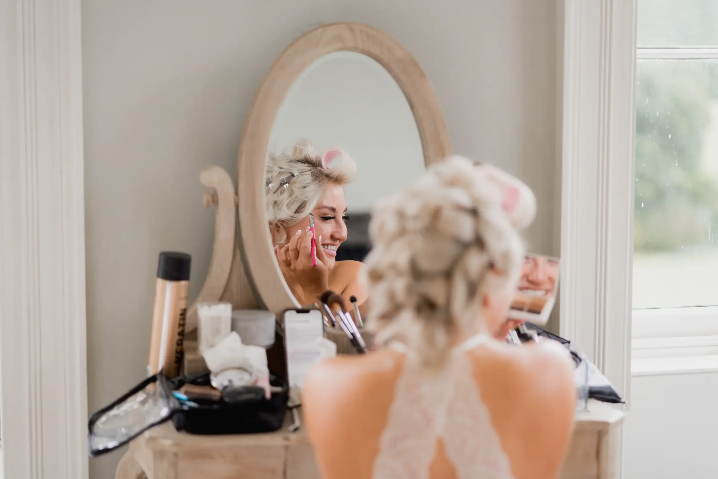 Woman smiling with hair rollers in her hair, applying makeup in front of a mirror on a dressing table.
