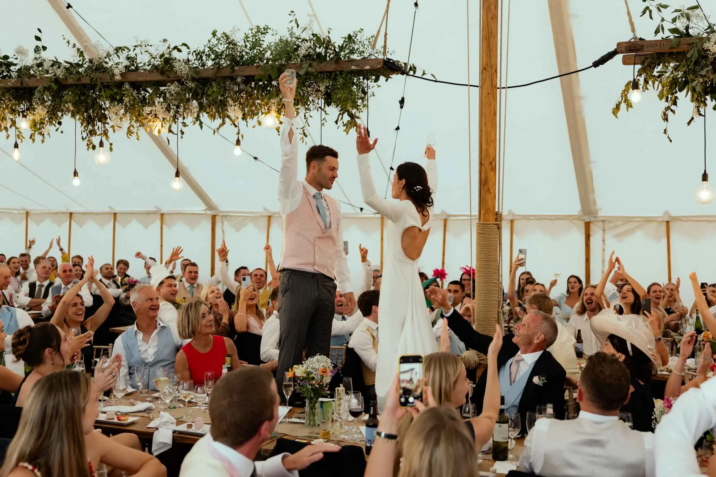 Bride and groom dancing on a table at wedding reception with guests cheering and taking photos inside a decorated tent.