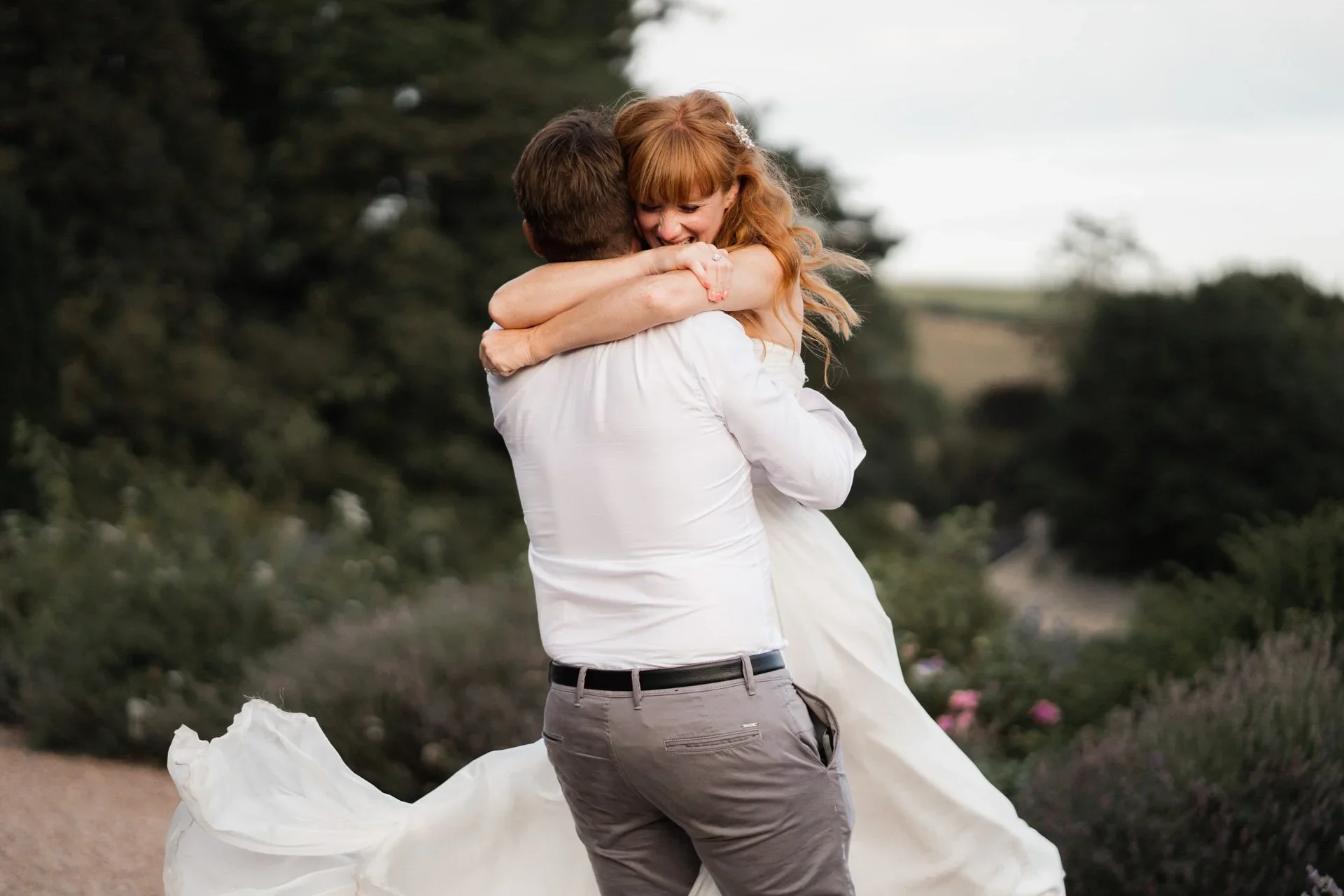 A man and woman embrace outdoors, the woman in a white dress, the man in a white shirt and gray pants, with trees and shrubbery in the background.