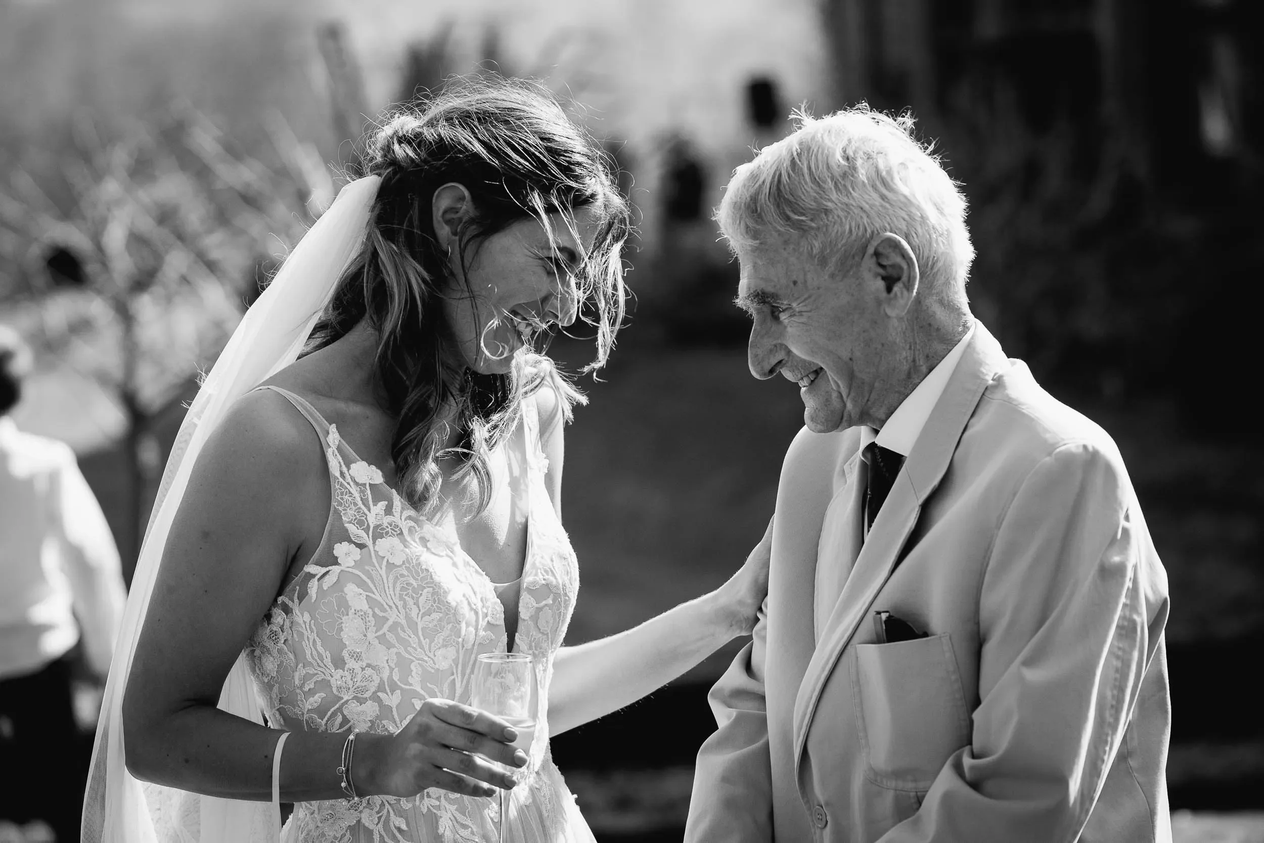 A bride in a lace wedding dress laughs with an elderly man dressed in a light-colored suit outdoors during a wedding celebration.