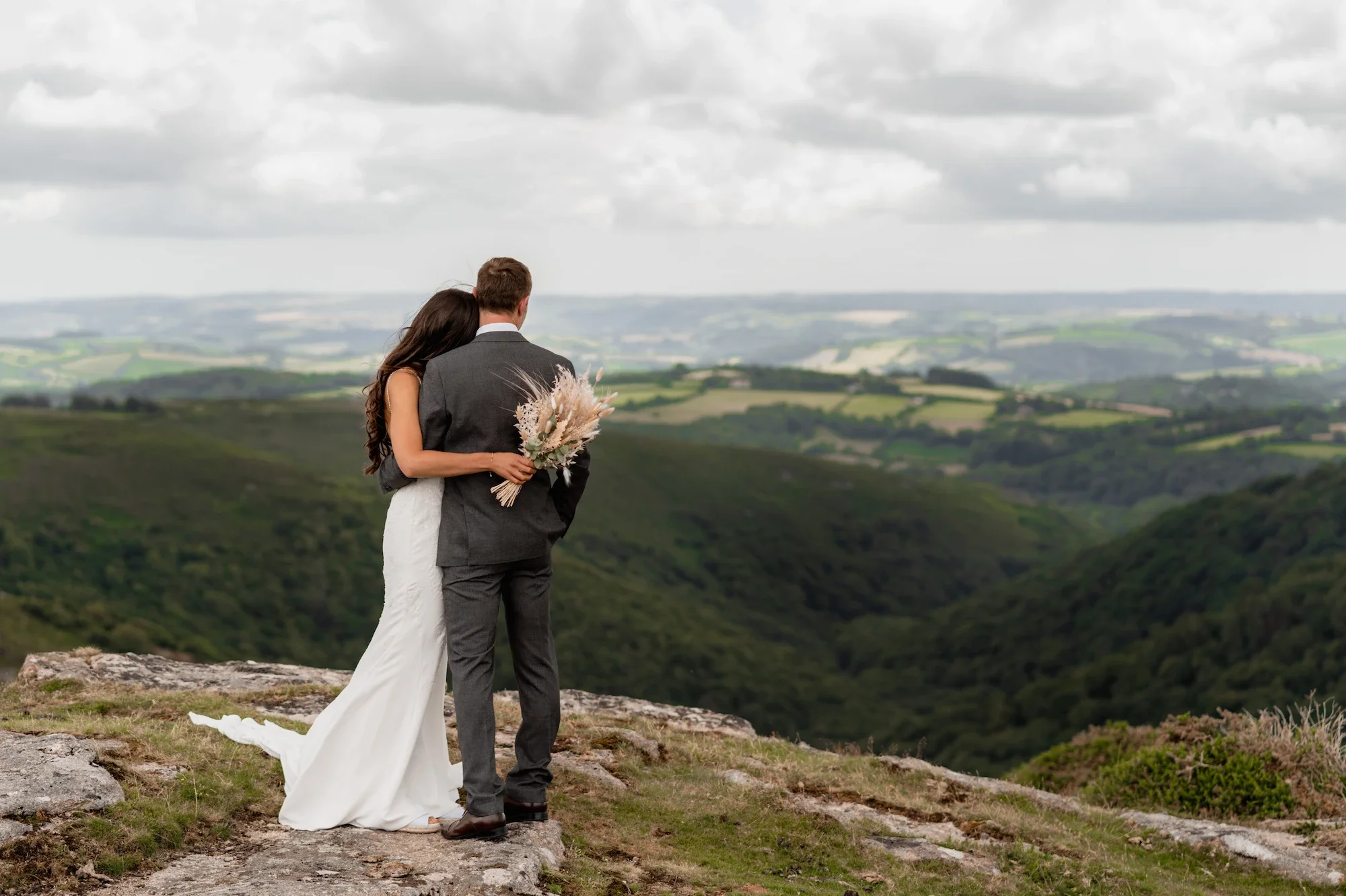 Wedding couple stands on rocky cliff overlooking green rolling hills, the bride in a white dress with long dark hair, the groom in a gray suit, holding a bouquet of pampas grass.