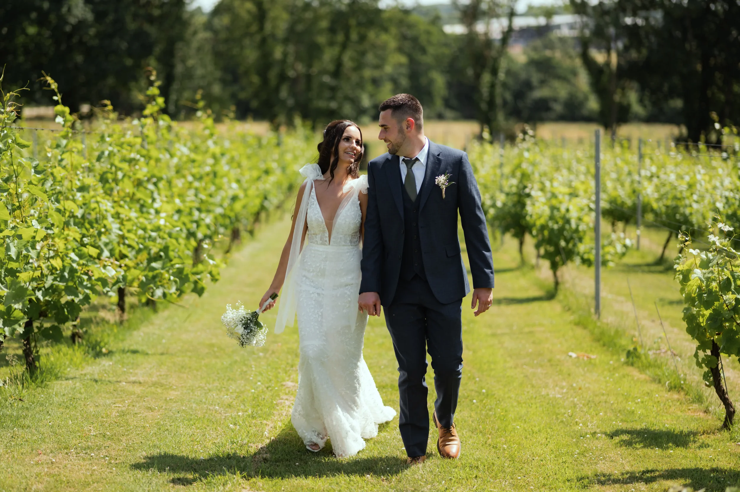 A bride and groom walking hand in hand through a vineyard on their wedding day, smiling at each other.