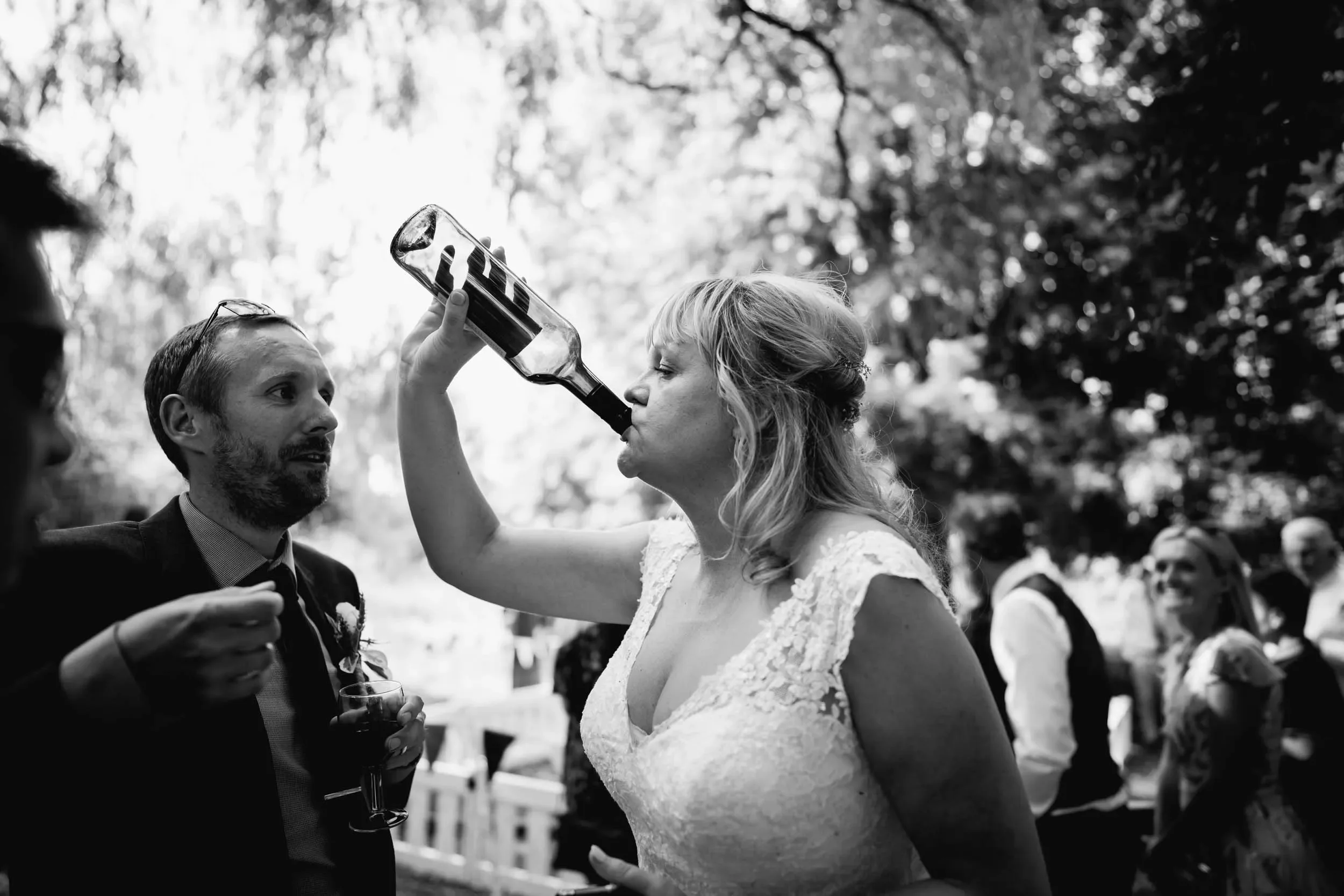 A woman in a wedding dress drinking beer from a bottle at an outdoor wedding reception, surrounded by other people.