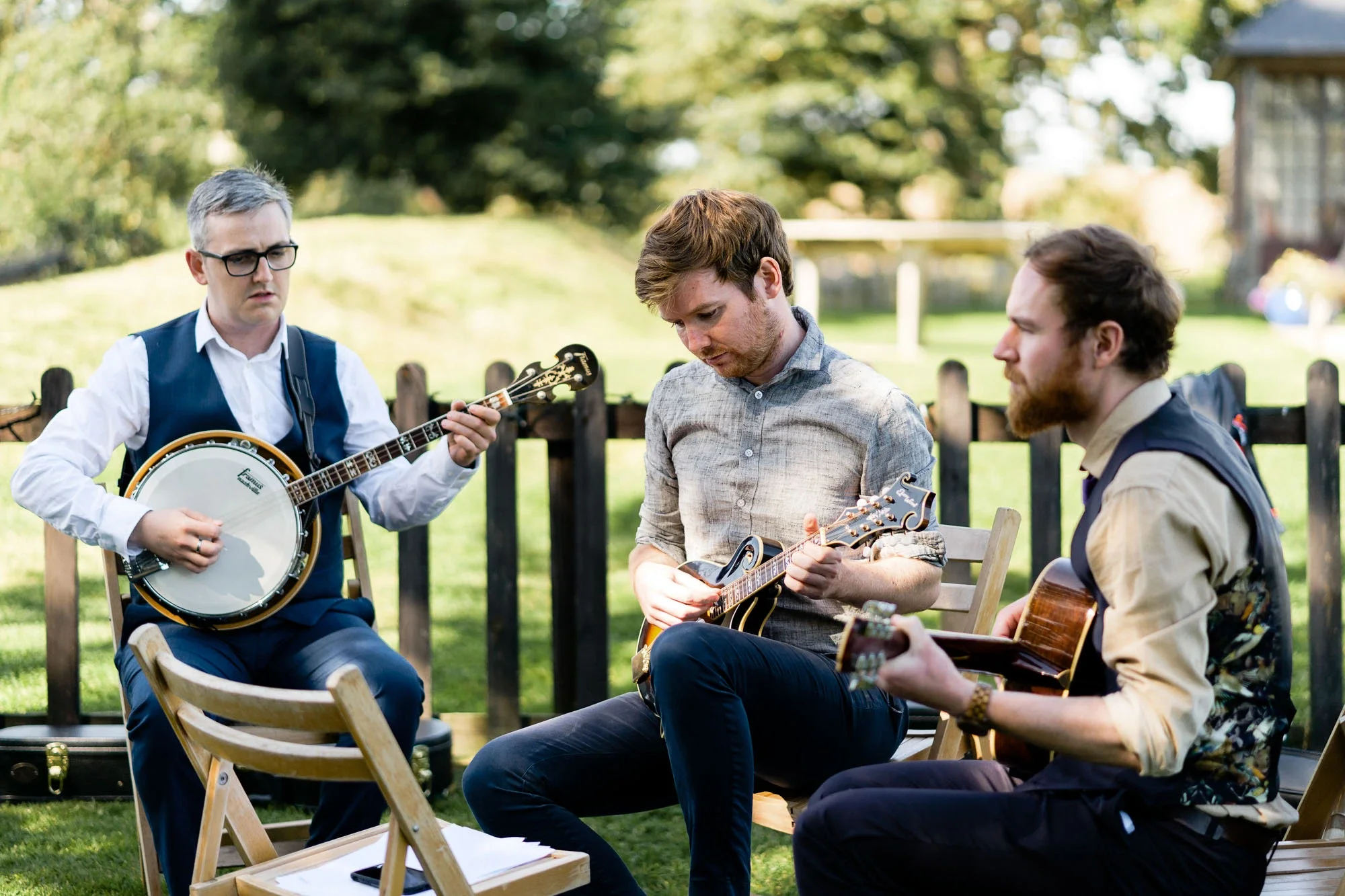 Three men playing musical instruments outdoors on a grassy lawn with trees and a wooden fence in the background. One man is playing a banjo, one is playing a mandolin, and the third is playing an acoustic guitar.