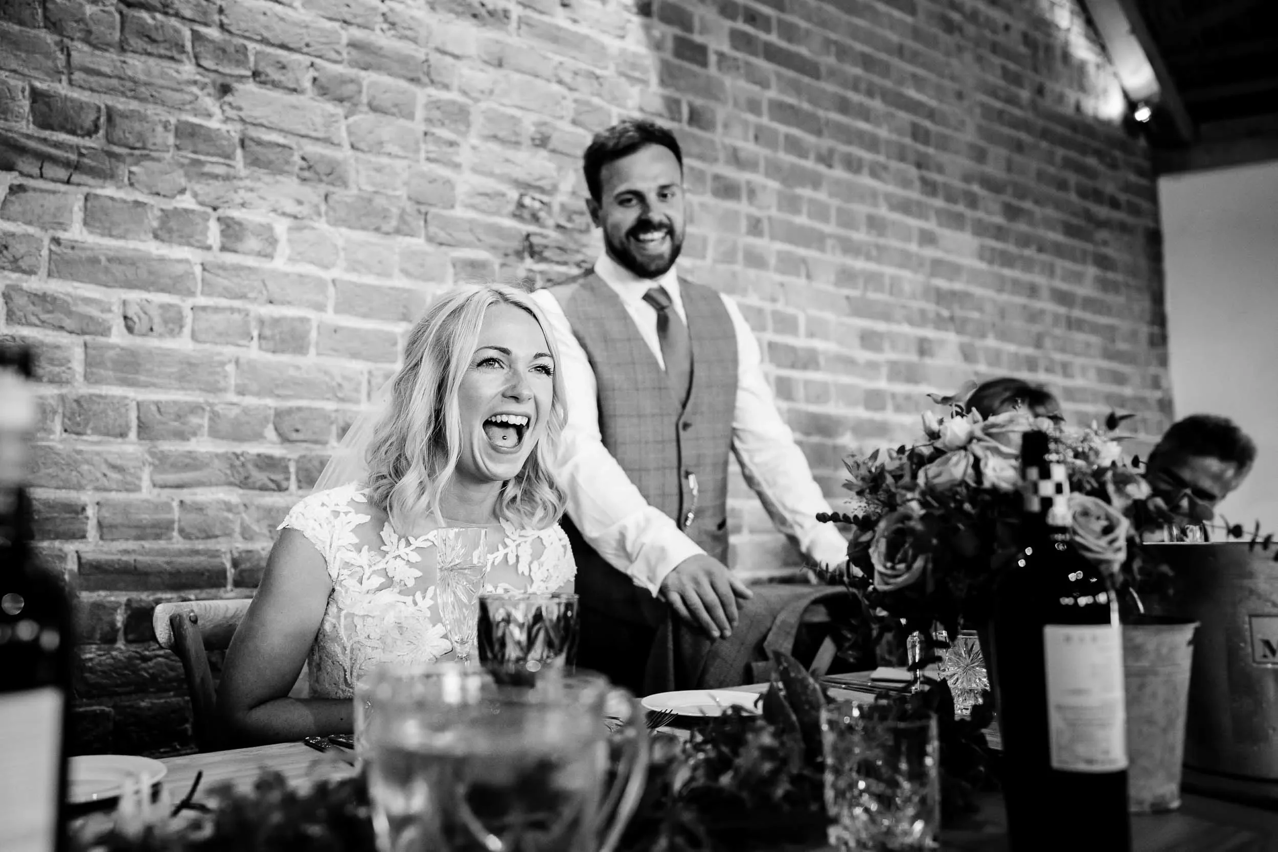 Black and white photo of a woman laughing and a man standing behind her at a celebration, with a brick wall in the background and table with flowers and drinks in the foreground.