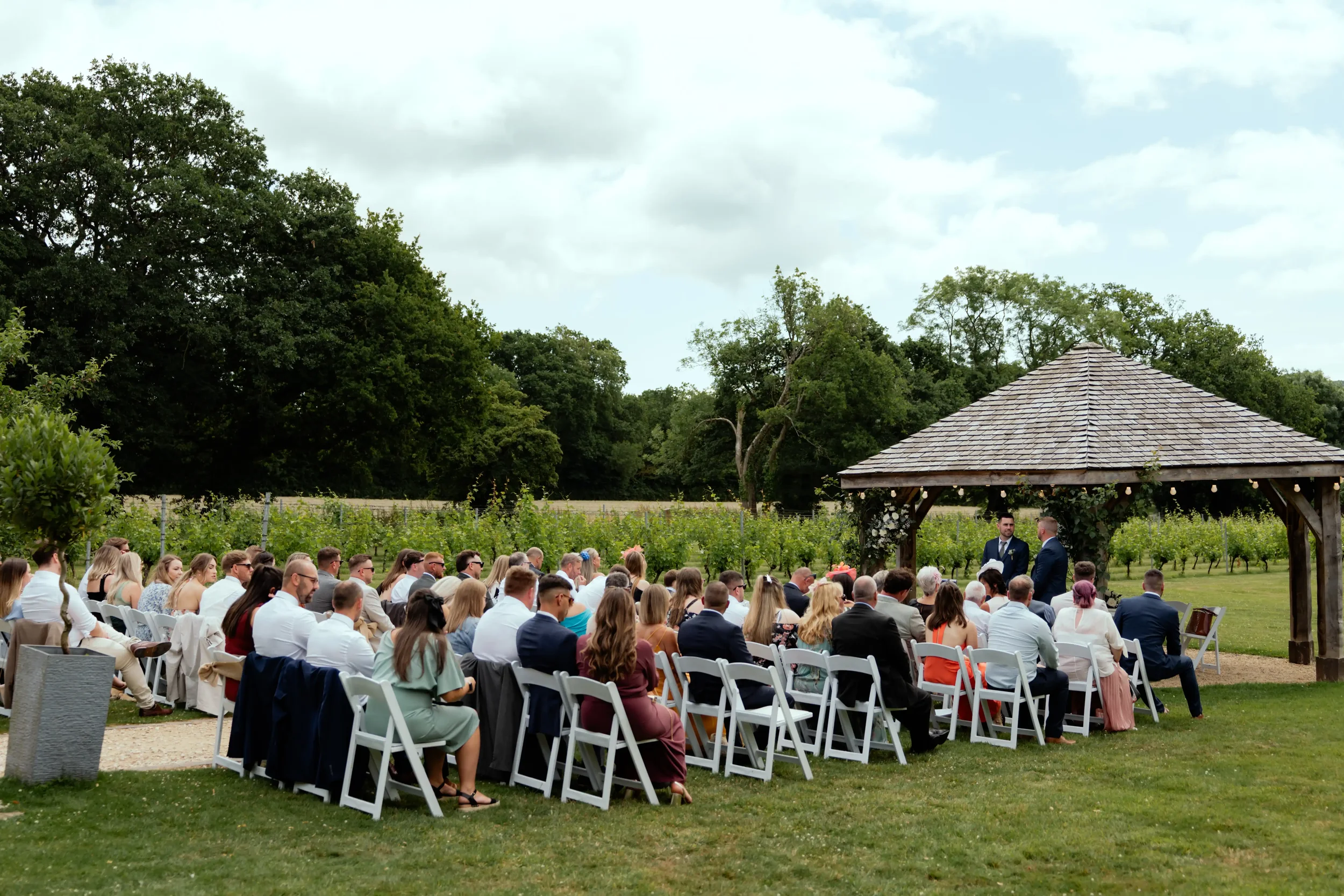 Outdoor wedding ceremony with many guests seated on white chairs under a gazebo in a lush green vineyard setting.