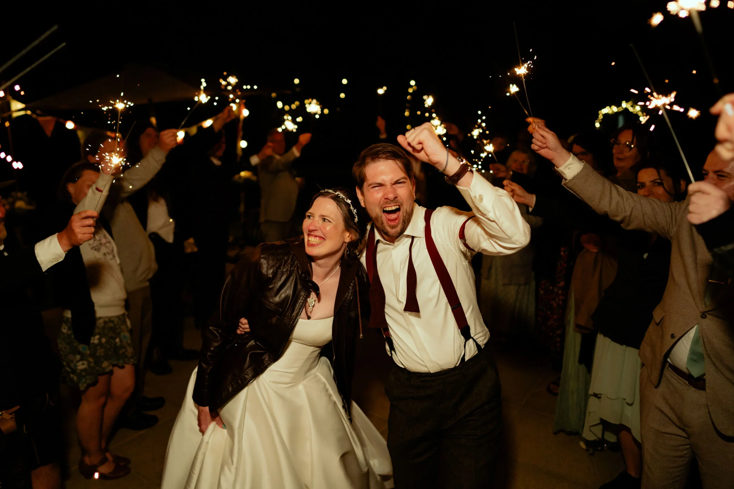 People celebrating at a nighttime event with sparklers, including a woman in a white dress and leather jacket, and a man in a white shirt with suspenders, involved in a lively celebration.