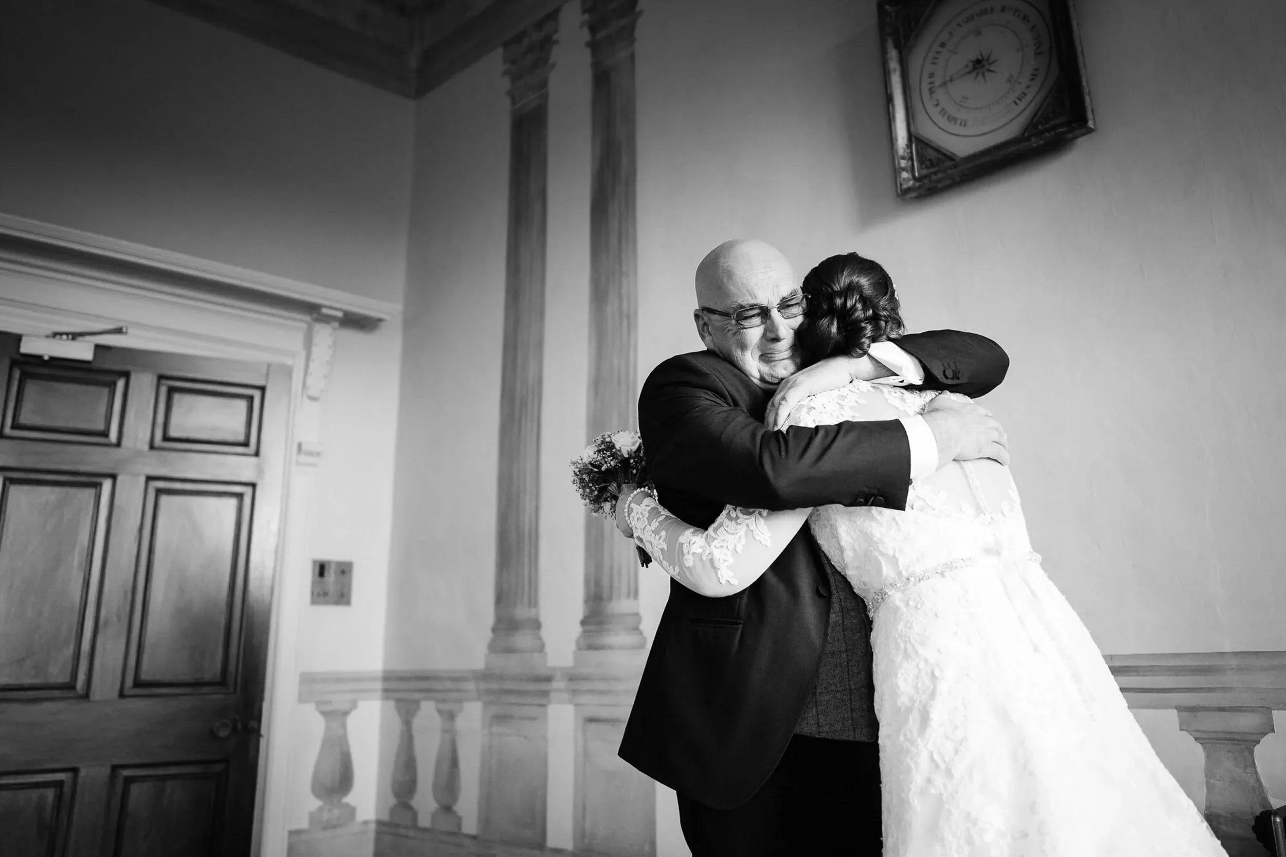 An elderly man in a suit and a woman in a wedding dress hugging in a room with wooden architectural details and a clock on the wall.