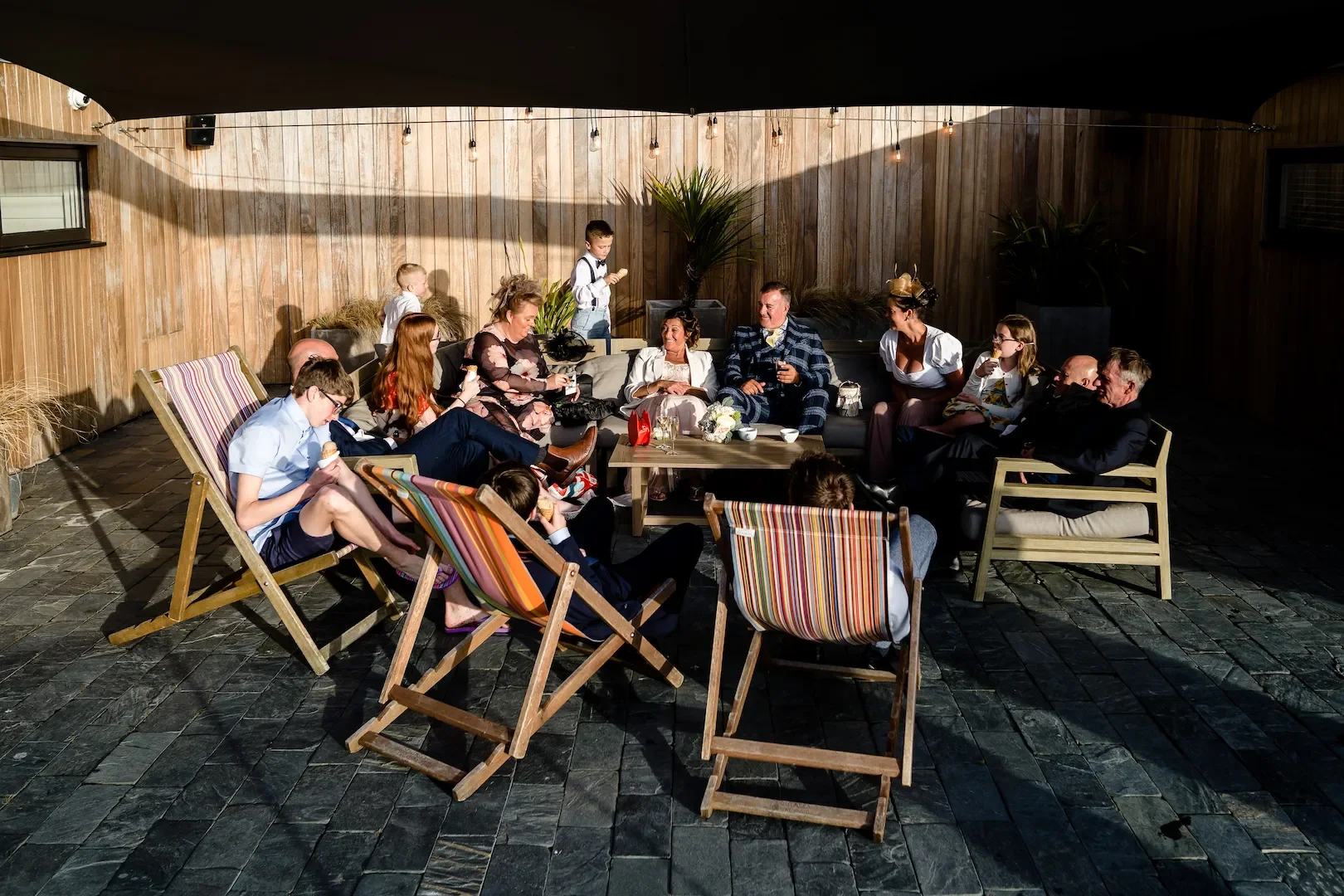People enjoying an outdoor gathering on a patio with wooden walls, some sitting on chairs and others on a sofa, with children and adults socializing and eating ice cream.