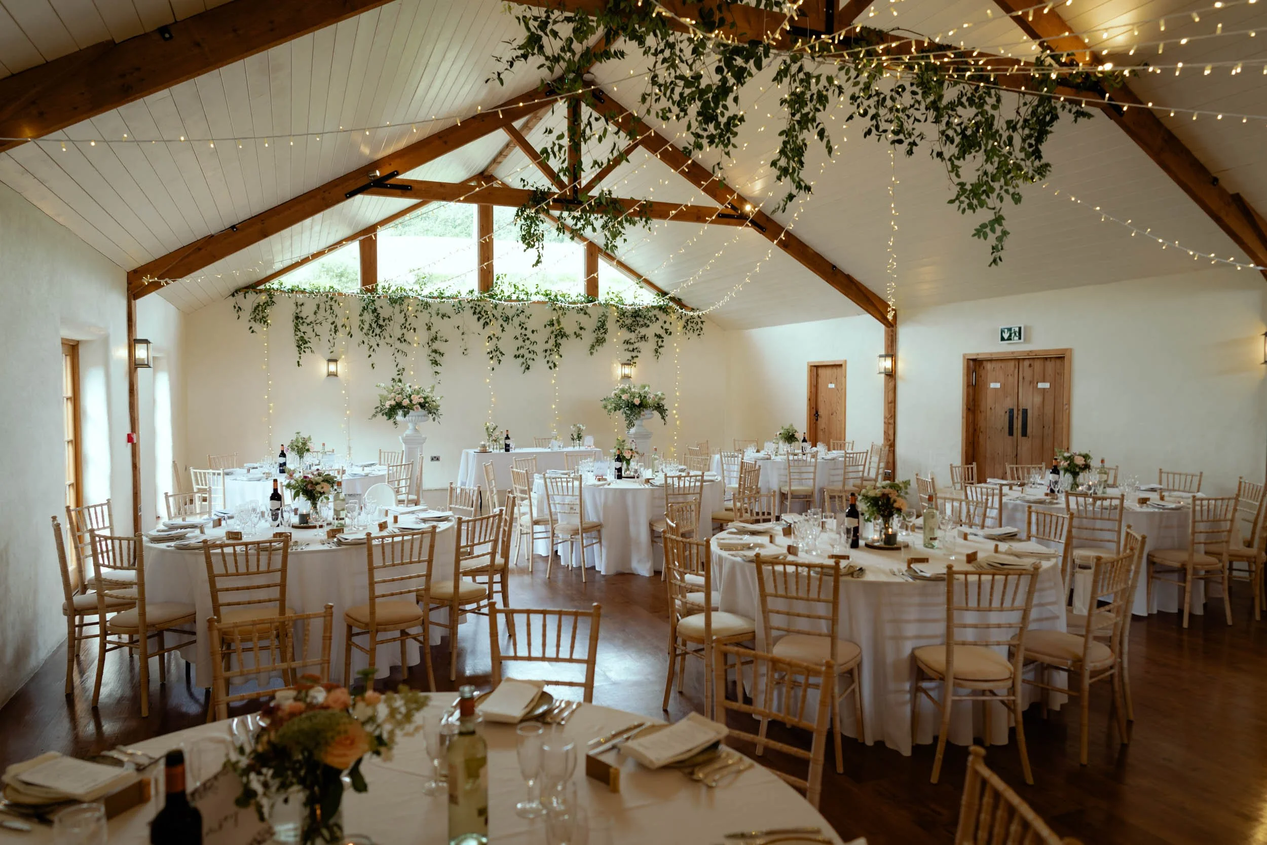 Elegant wedding reception setup in a decorated barn with round tables covered in white tablecloths, floral centerpieces, candles, and black wine bottles. The barn features wooden beams, string lights, and greenery hanging from the ceiling, with large