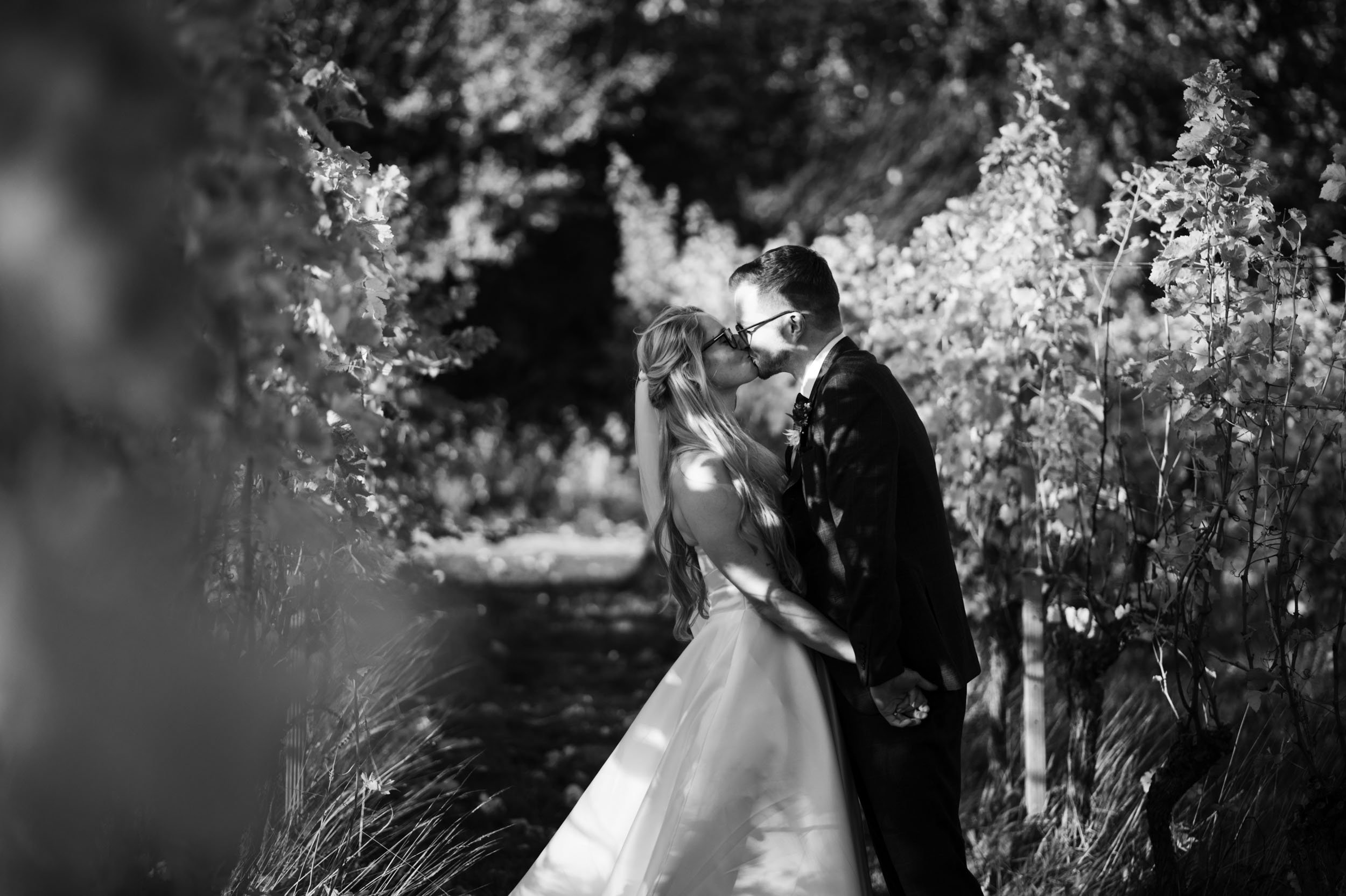 A black and white photograph of a bride and groom kissing outdoors, surrounded by trees and foliage, holding hands, with sunlight filtering through the leaves.