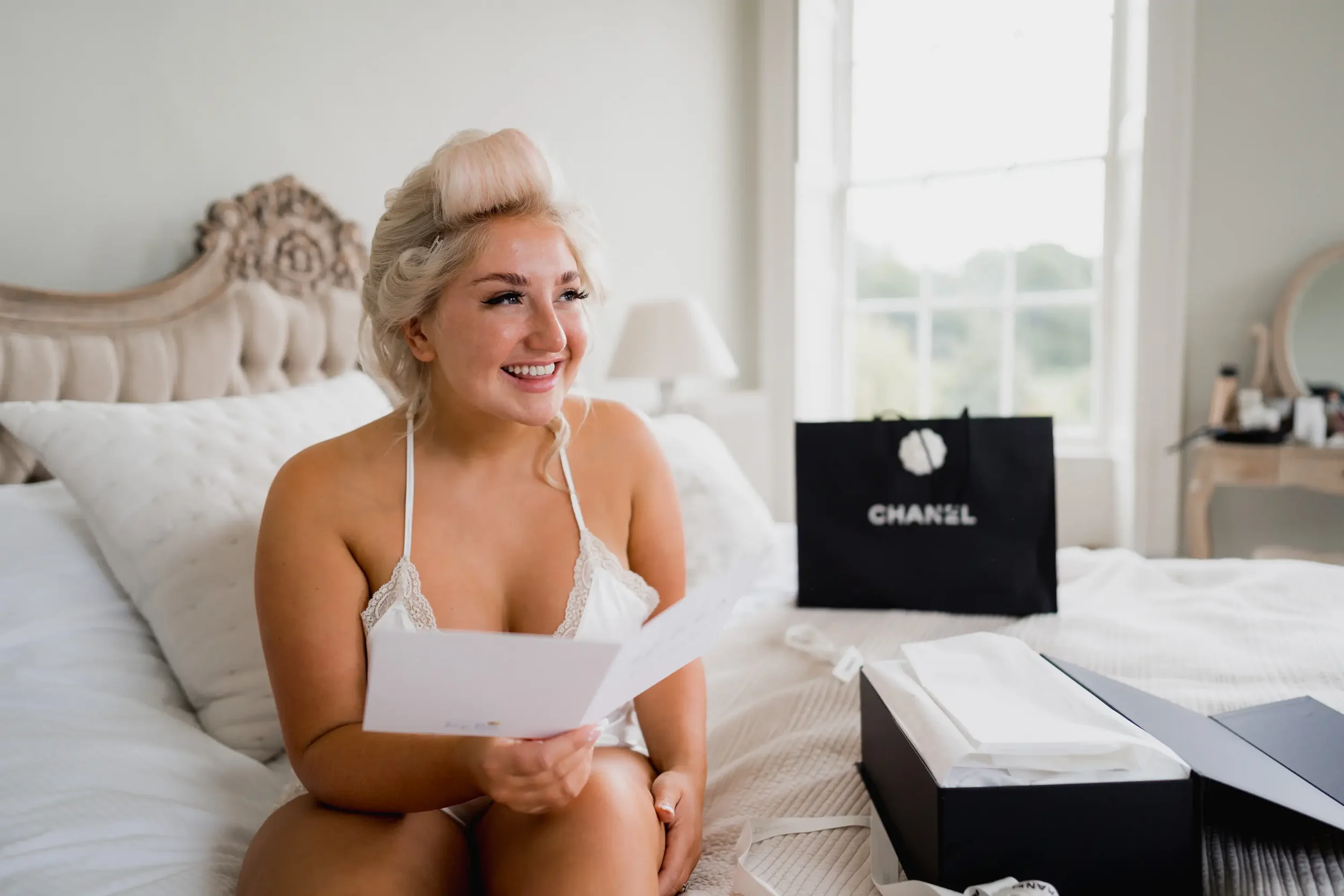 A woman with blonde hair sitting on a bed, smiling while looking at a letter. There is a Chanel shopping bag and tissue box on the bed.