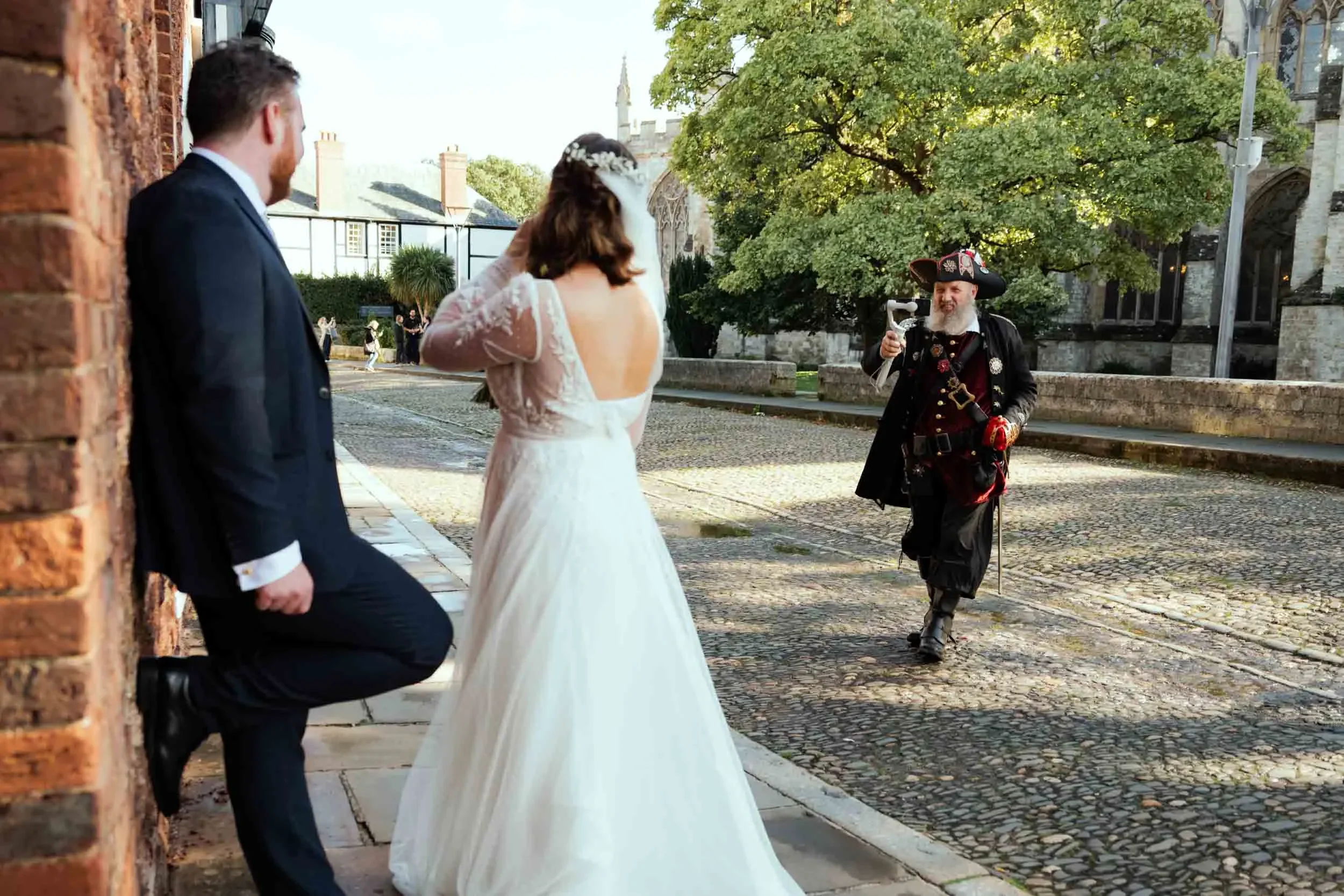 A wedding scene with a bride in a white gown and a groom in a dark suit, standing outside on cobblestone pavement. An older man dressed as a pirate is approaching them, holding a horn as if about to play it.