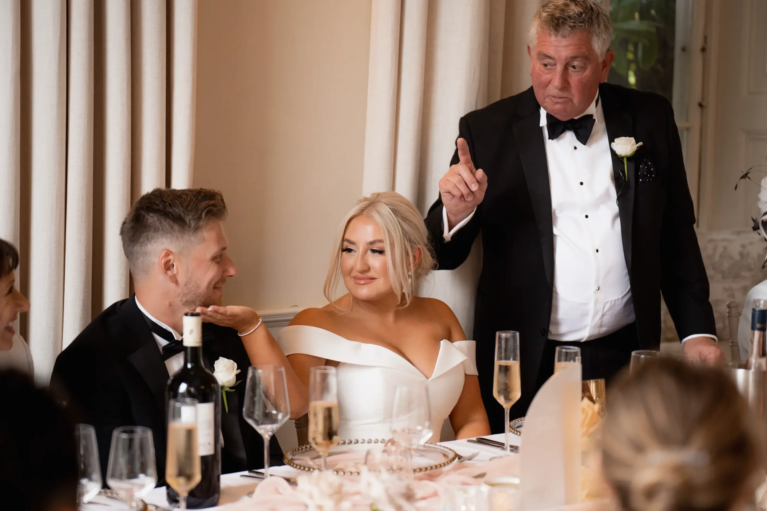A wedding reception scene with a bride, groom, and an older man, likely giving a speech. The bride has blonde hair and is wearing an off-shoulder white dress, the groom has short light brown hair and is in a black tuxedo, and the older man is in a bl