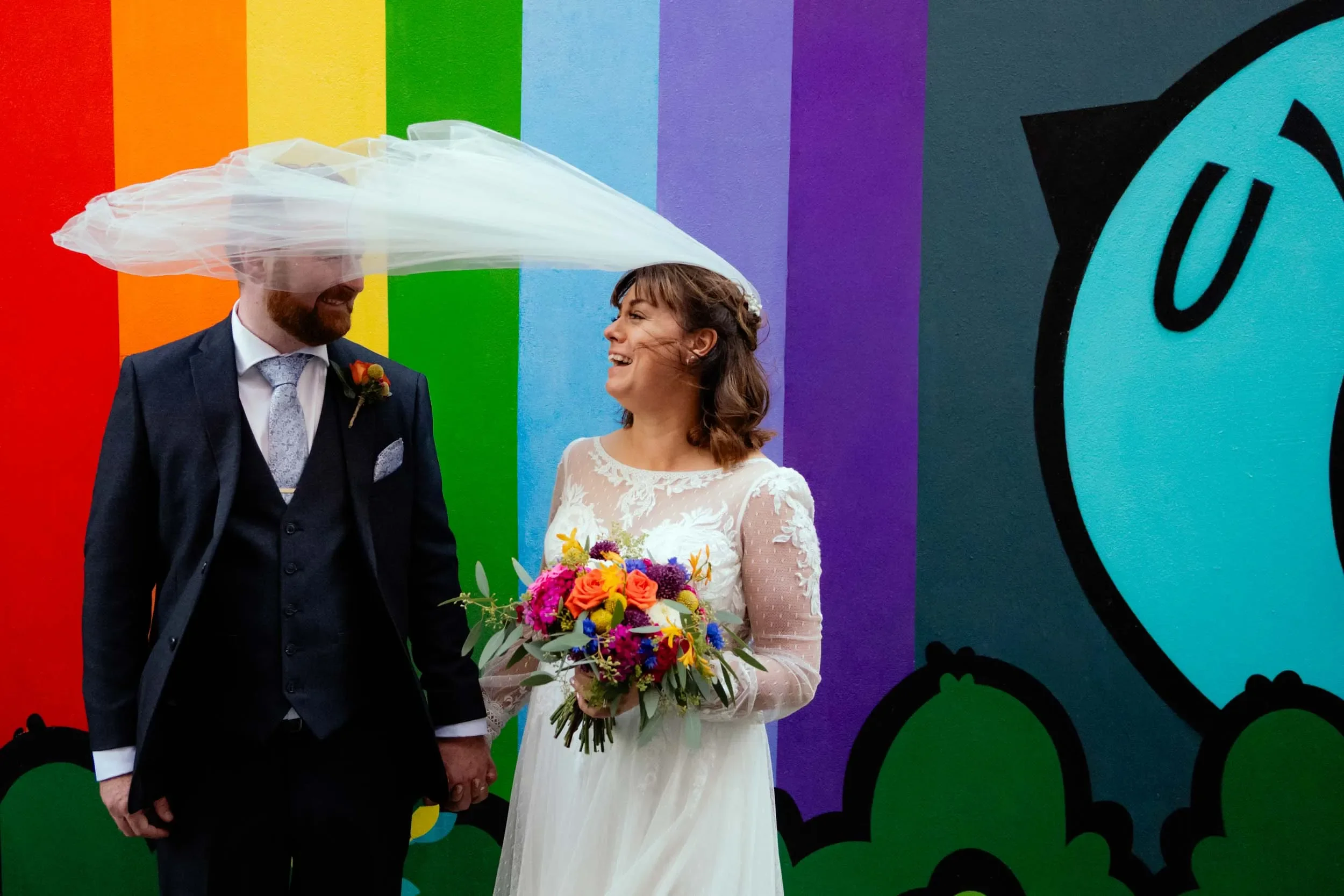 A bride and groom holding hands at their wedding, standing in front of a colorful wall with rainbow stripes. The bride is smiling and holding a bouquet of vibrant flowers, while the groom is wearing a suit with a boutonniere and has a sheer veil over