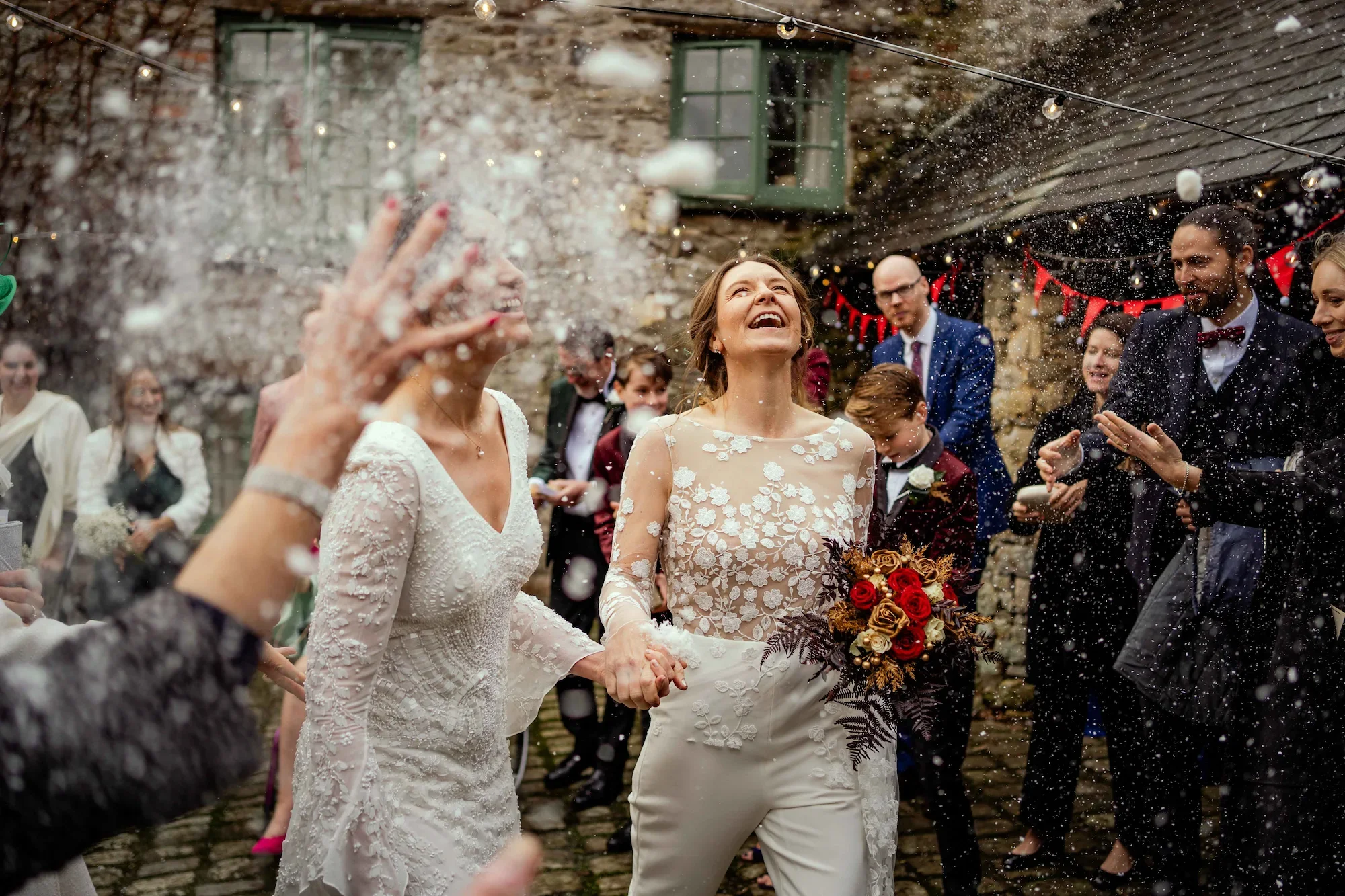 A bride and a woman, possibly her mother, celebrating outdoors during a wedding with guests throwing rice or confetti around them.