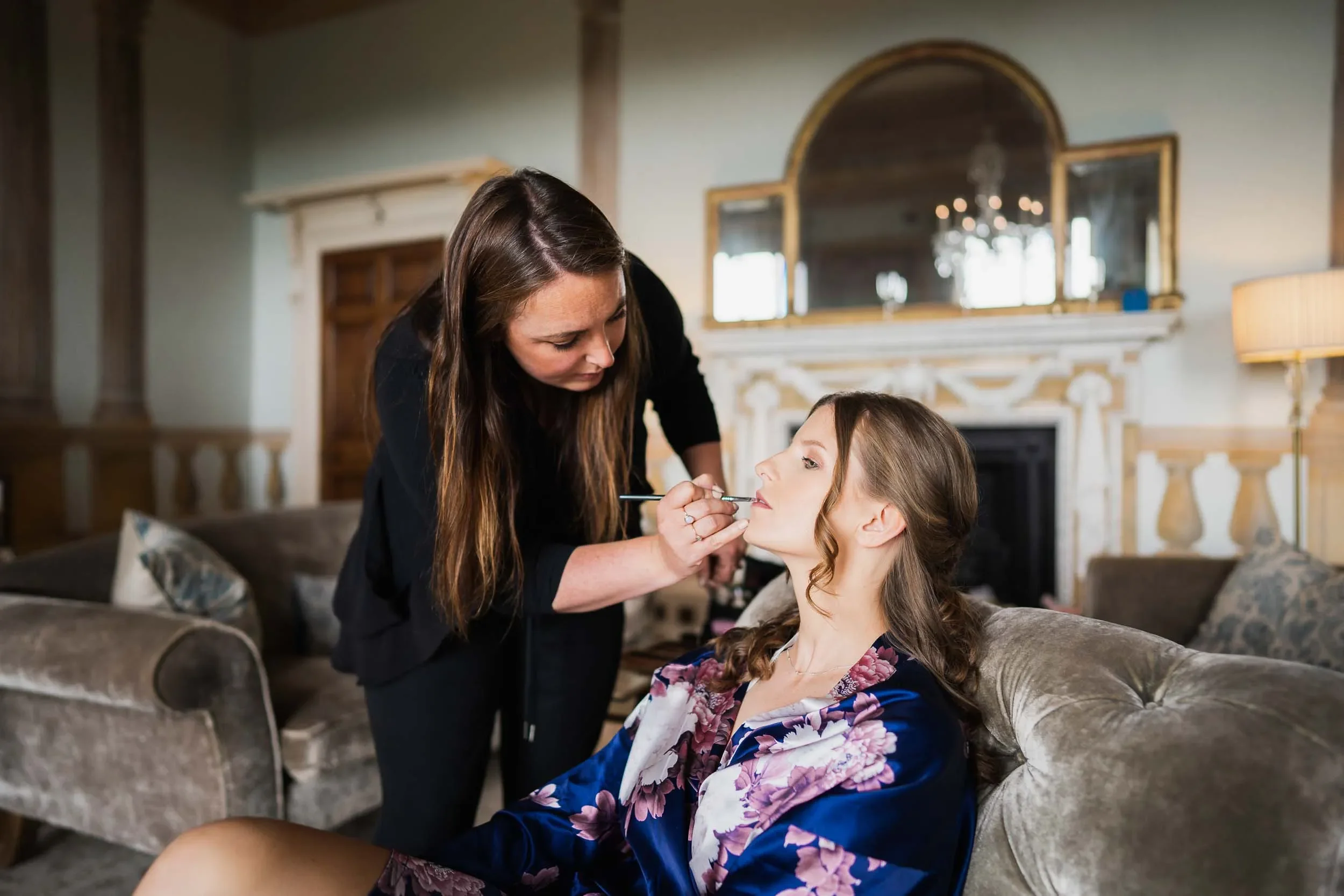 A makeup artist applying lipstick to a woman sitting on a sofa in a well-decorated living room.