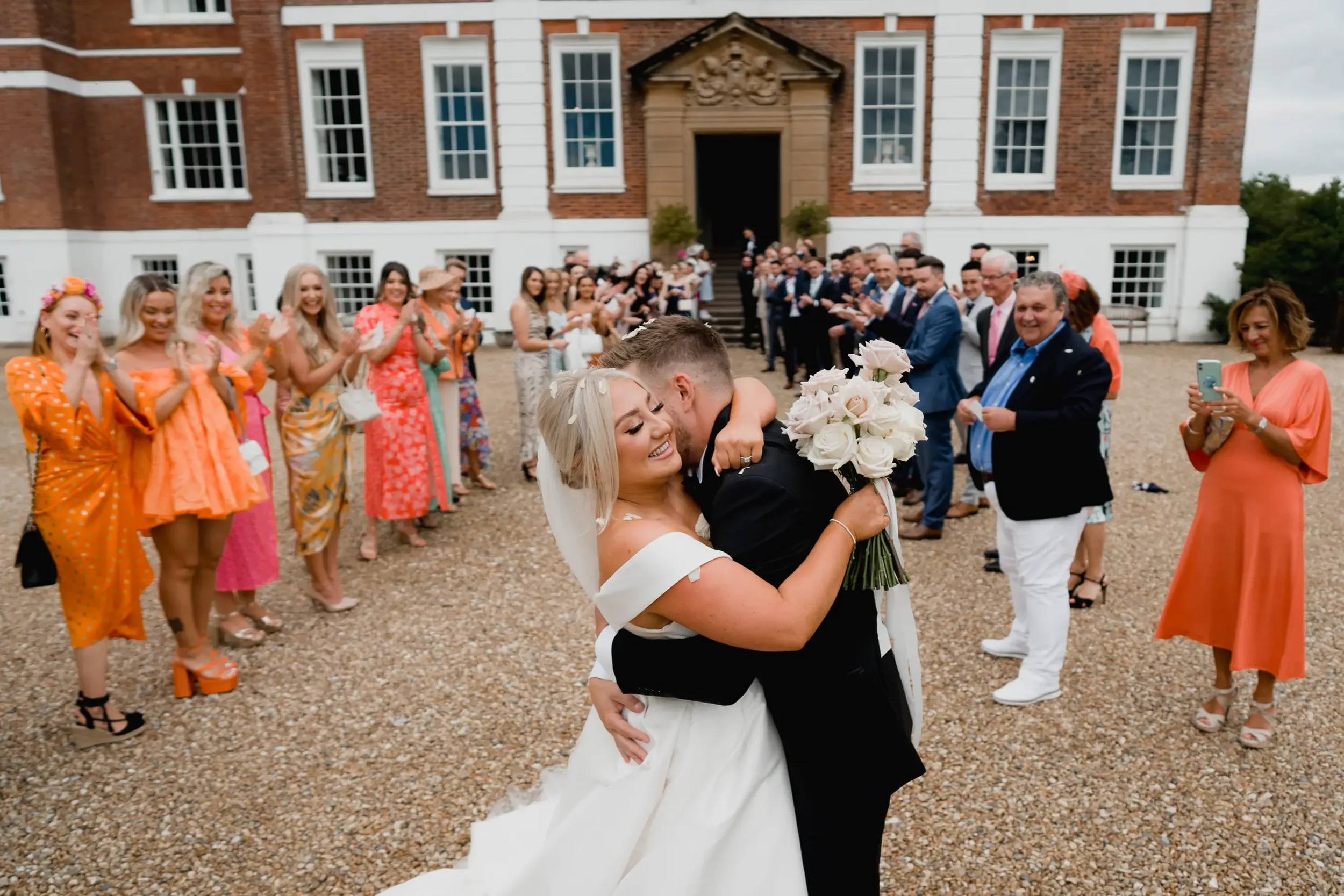 A bride and groom hugging and smiling in front of guests at a wedding in front of a large brick and white building with an open doorway, with guests clapping and taking photos.