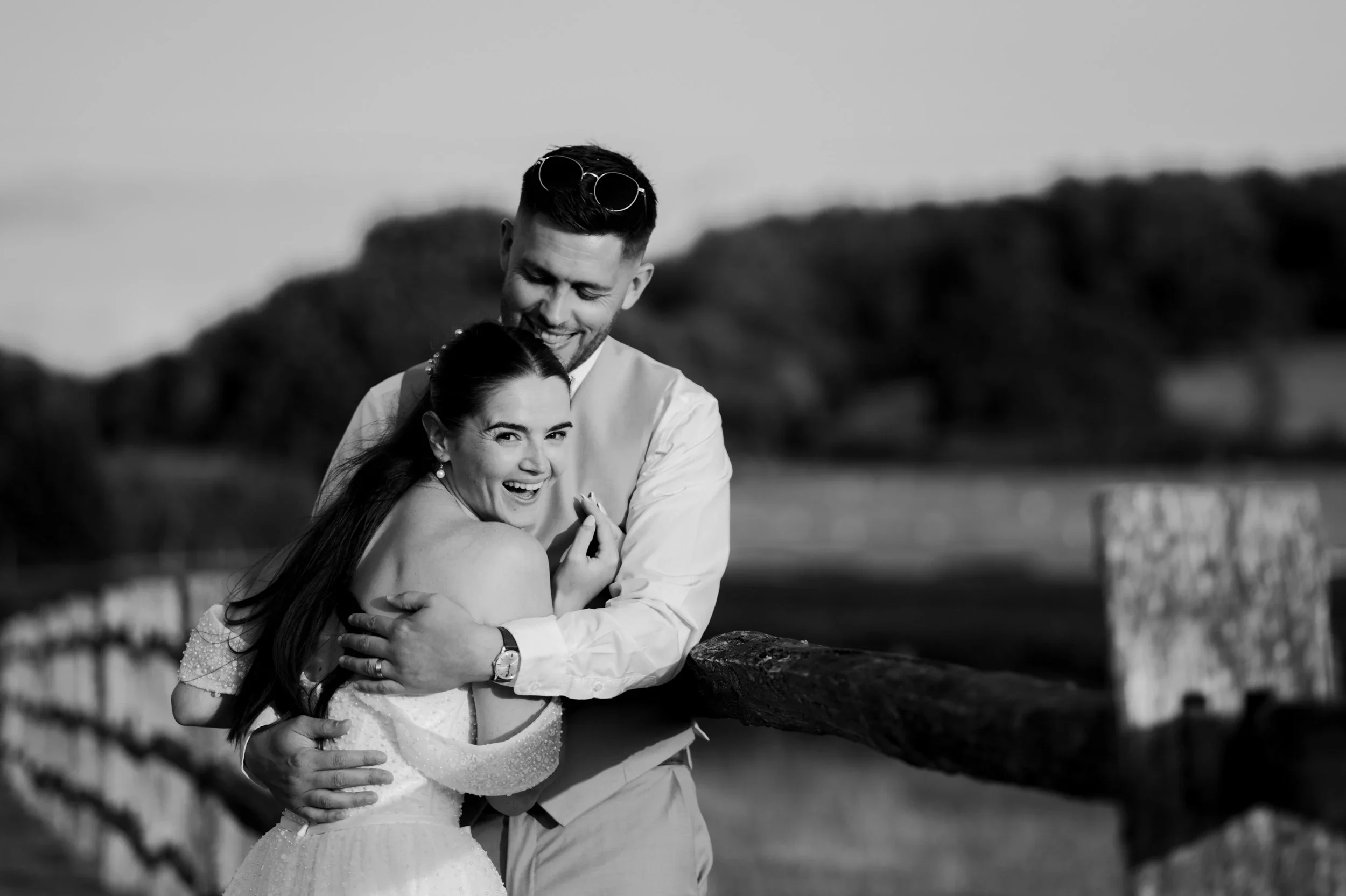 A black and white photo of a smiling couple embracing outdoors near a wooden fence, with blurred landscape and distant trees in the background.