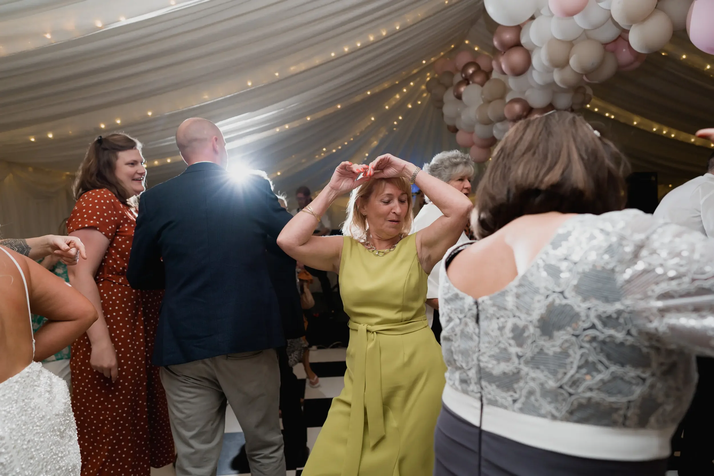 People dancing at a celebration under a decorated tent with balloon arrangements and string lights.