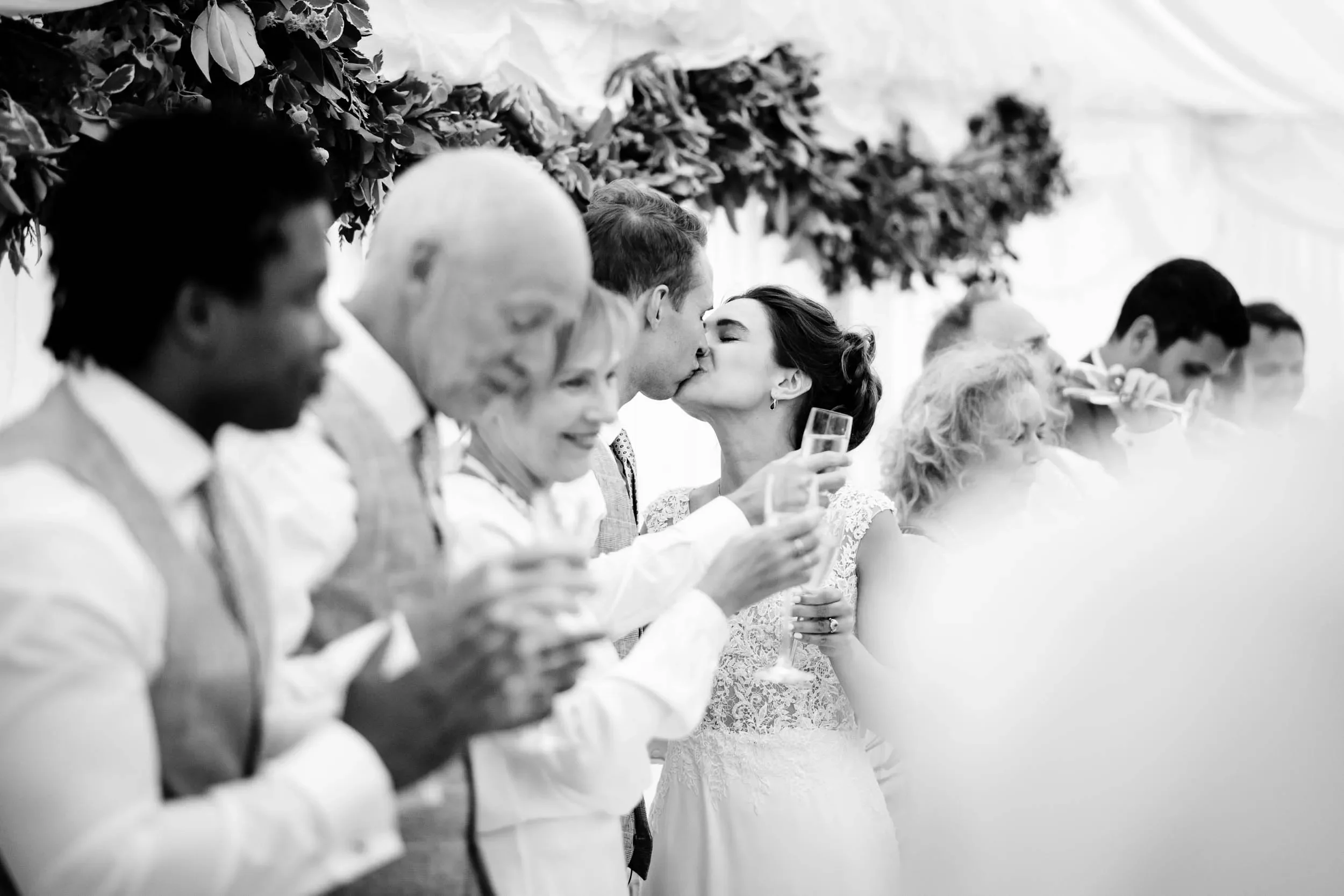 Black and white photo of a wedding celebration with a bride and groom kissing, surrounded by guests holding glasses, at a festive event.