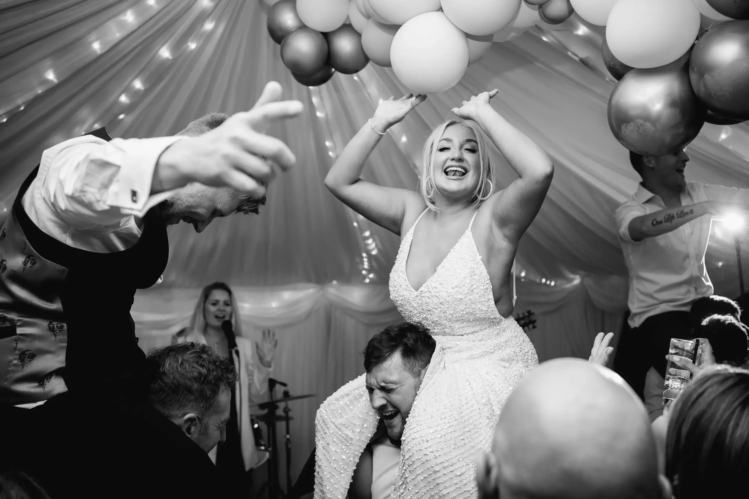 A bride and groom celebrating at a wedding reception, with the bride smiling and raising her hands while sitting on the groom's shoulders, surrounded by friends and family in a decorated tent with balloons.
