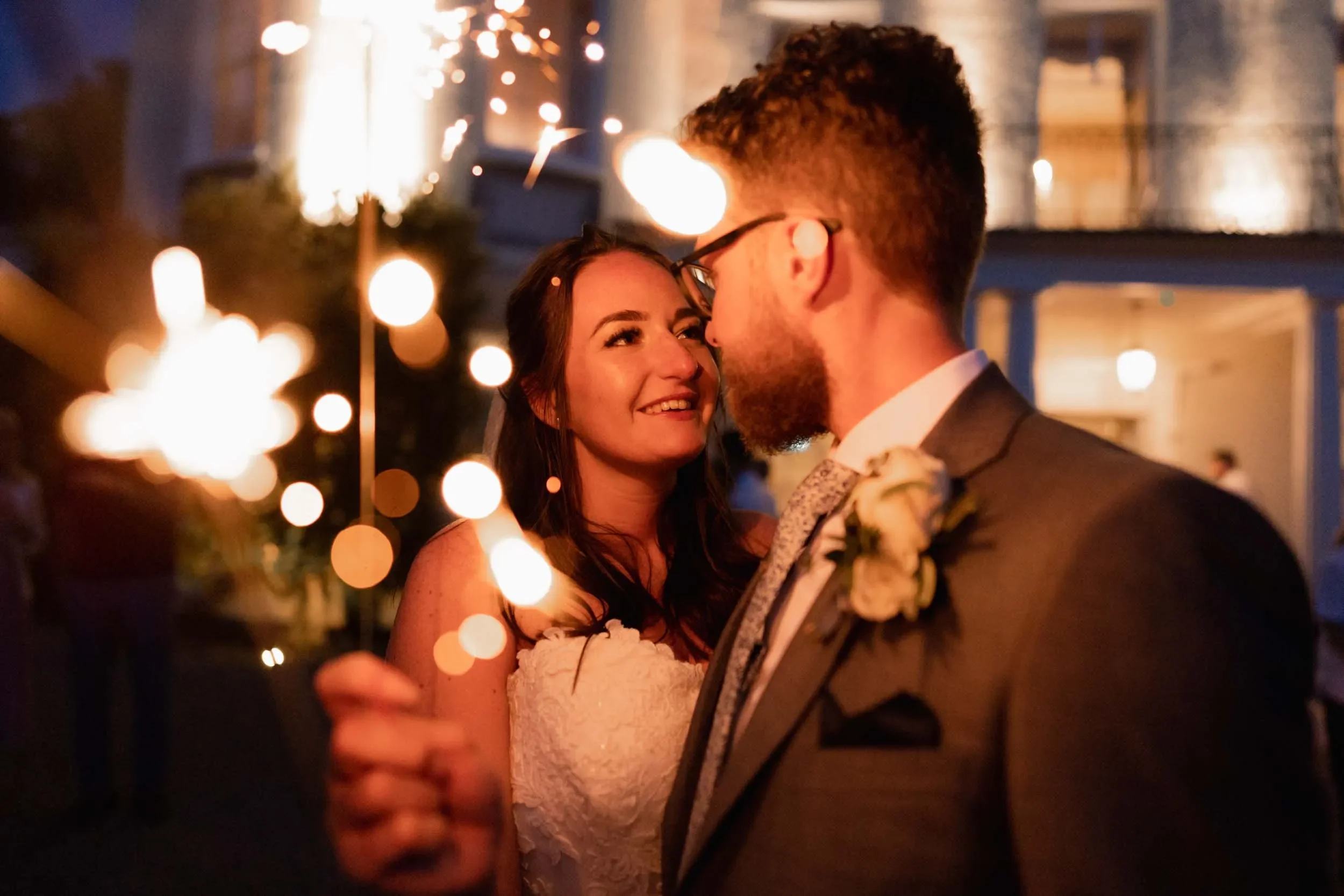 A newlywed couple at their wedding reception at night, holding sparklers and sharing a loving gaze amid warm, bokeh lights in the background.