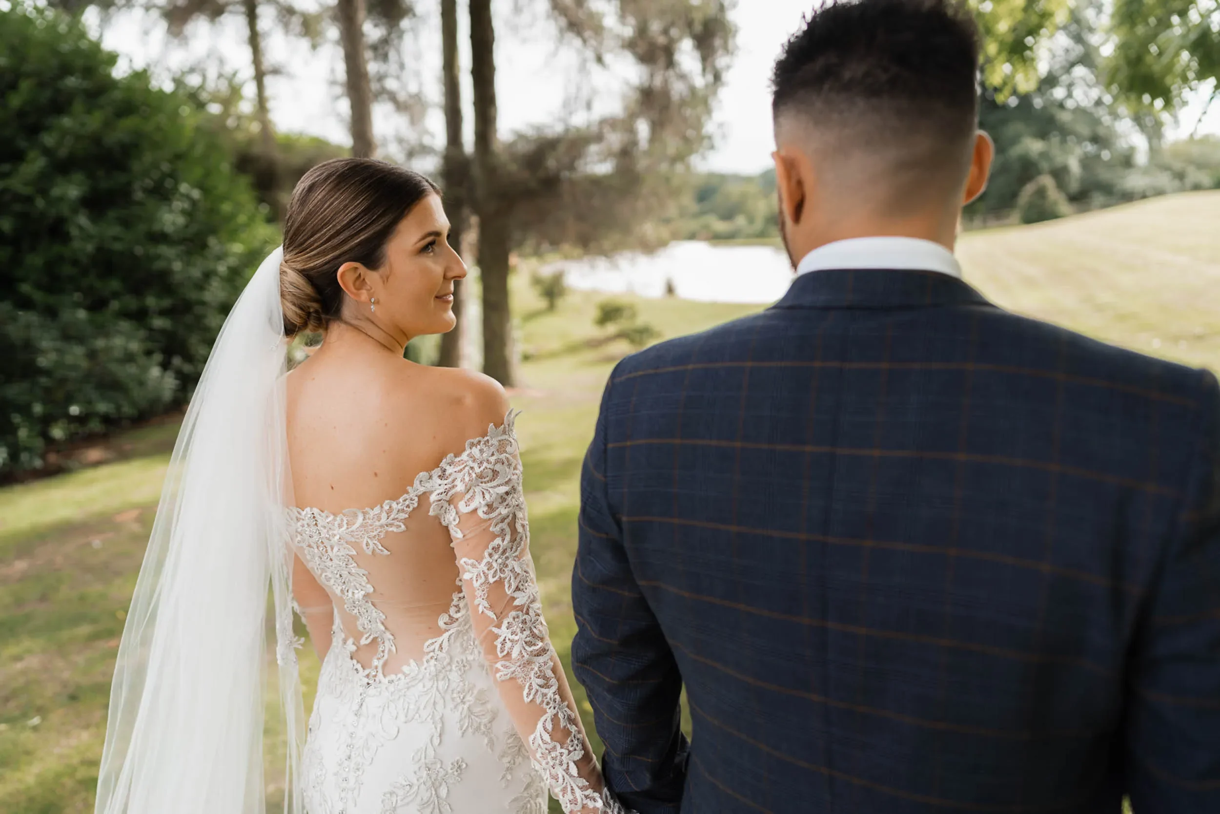 A bride and groom stand outdoors on their wedding day, holding hands, with the bride smiling and looking at the groom, in a natural setting with trees and a lake in the background.