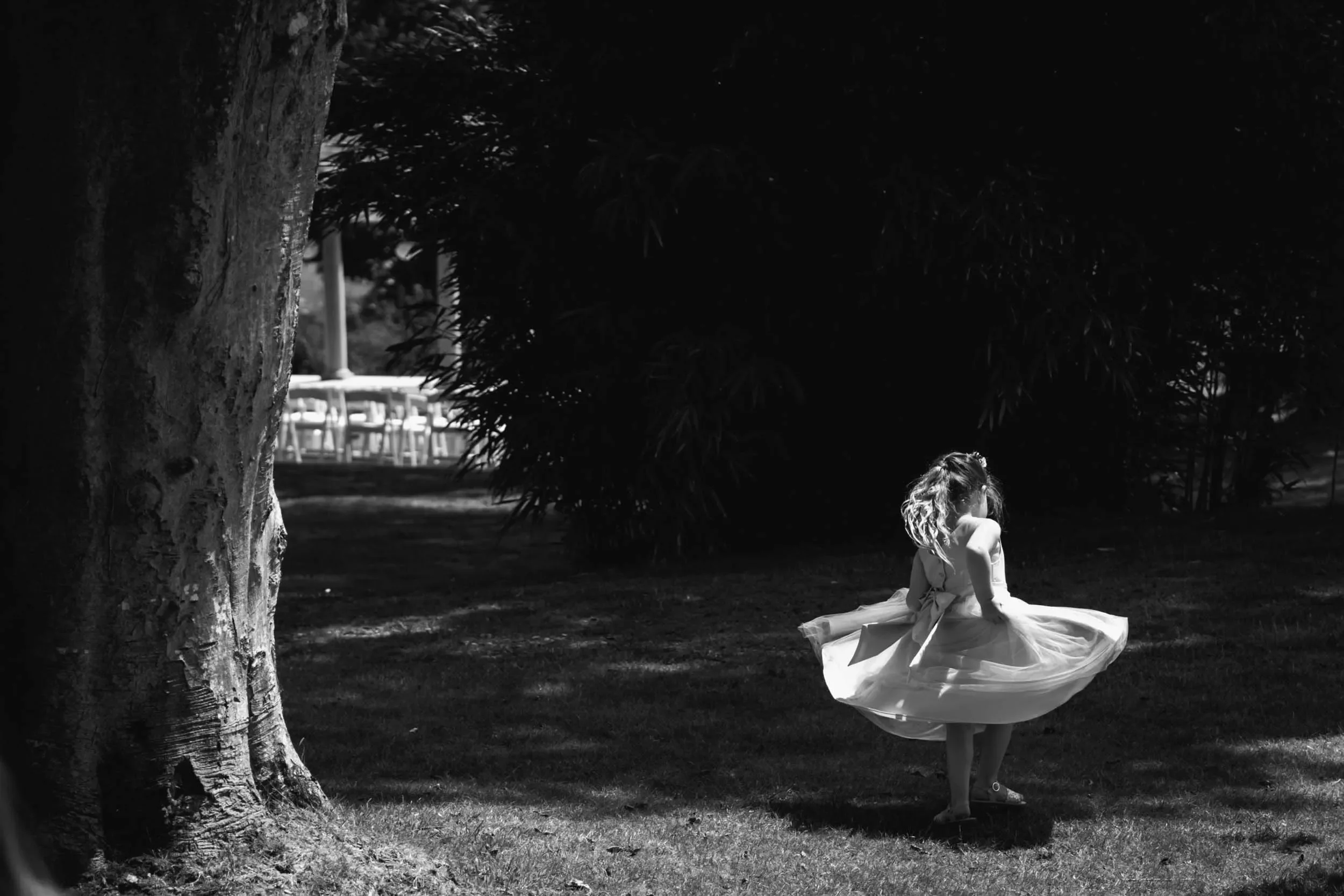 A young girl in a dress twirling outdoors in a park with a large tree and bushes, black-and-white photo.