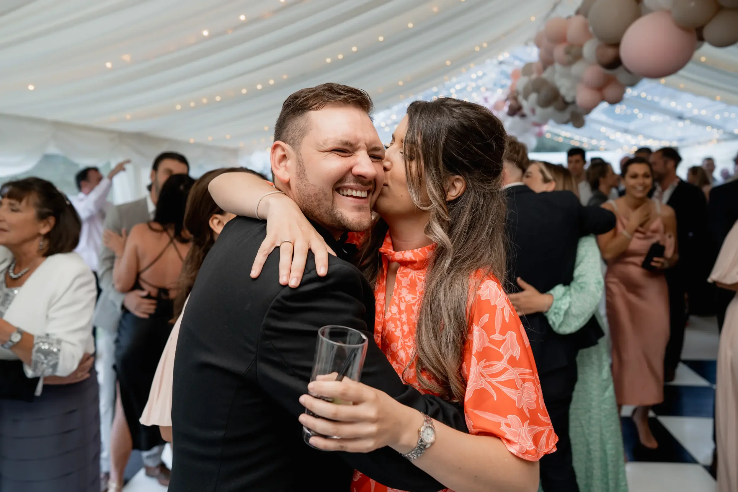 Couple hugging and smiling at a lively wedding reception with other guests dancing and socializing under a decorated tent.