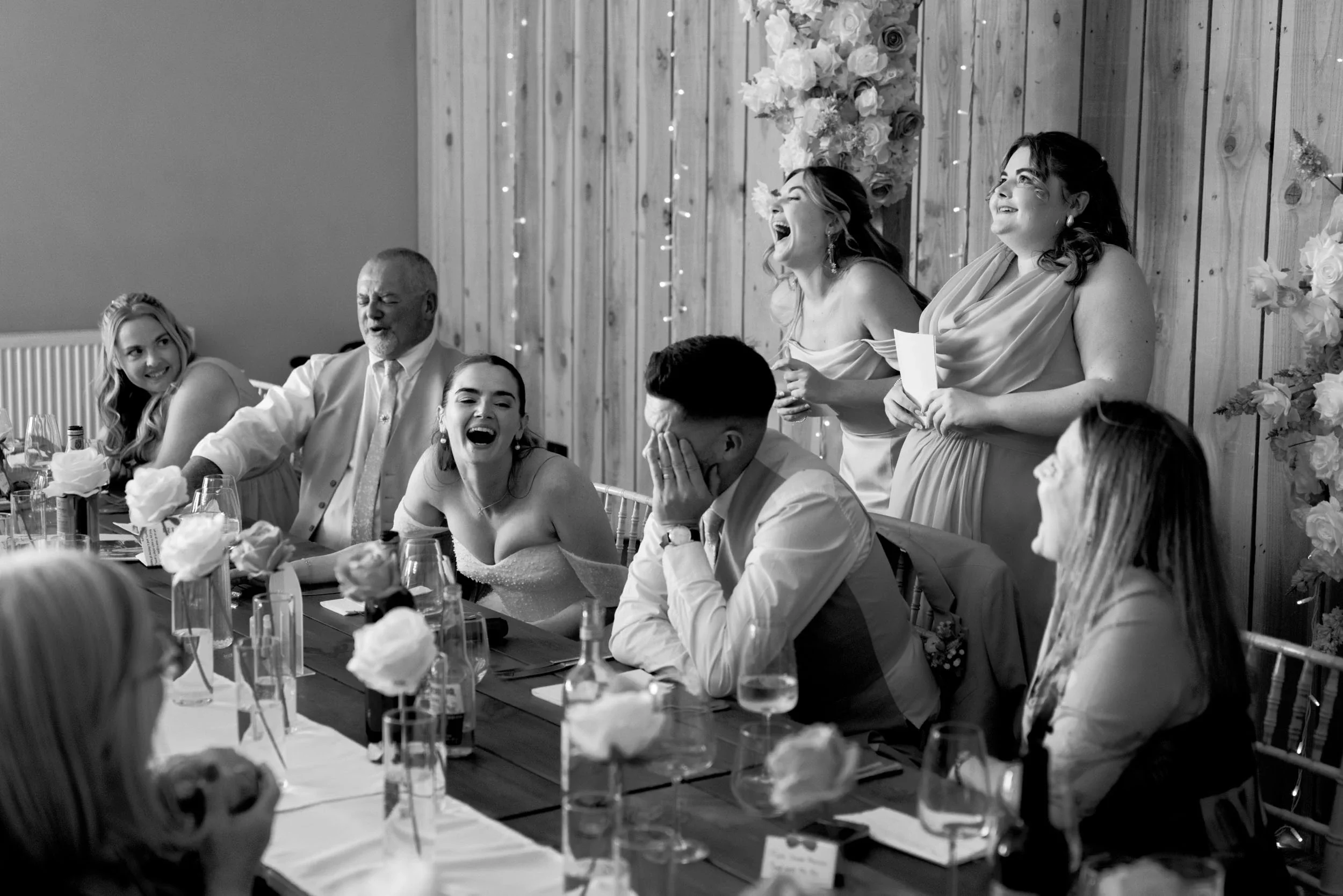 This black and white photo shows a wedding reception with guests seated at a long table, laughing and enjoying the moment. Behind them, a group of women is giving a toast, two of whom are standing and laughing, while one woman is holding a piece of p