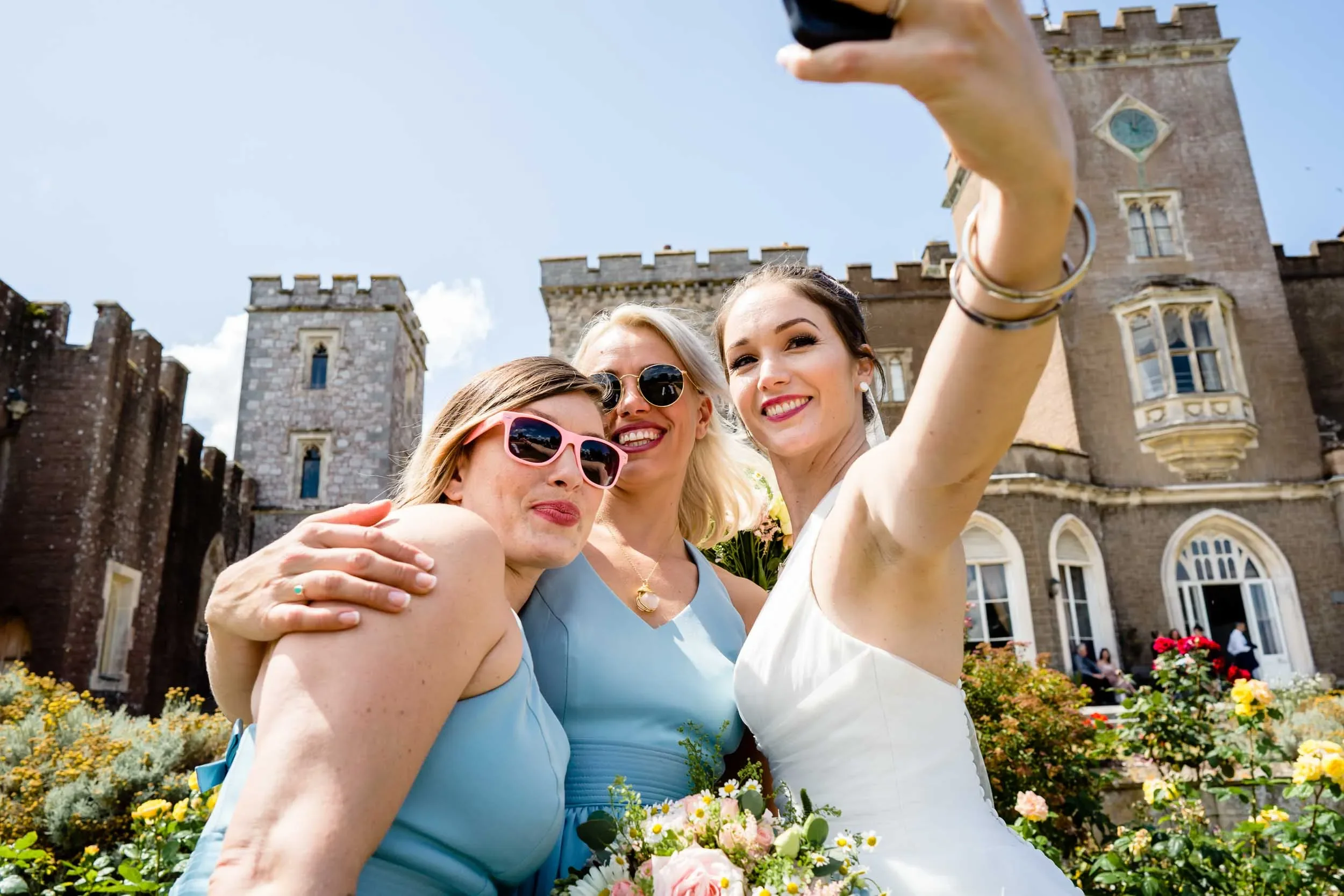 Three women taking a selfie in front of a castle on a sunny day, with flowers in the foreground. The woman on the right is wearing a white dress, likely a bride, and the other two women are dressed in light blue dresses with sunglasses.