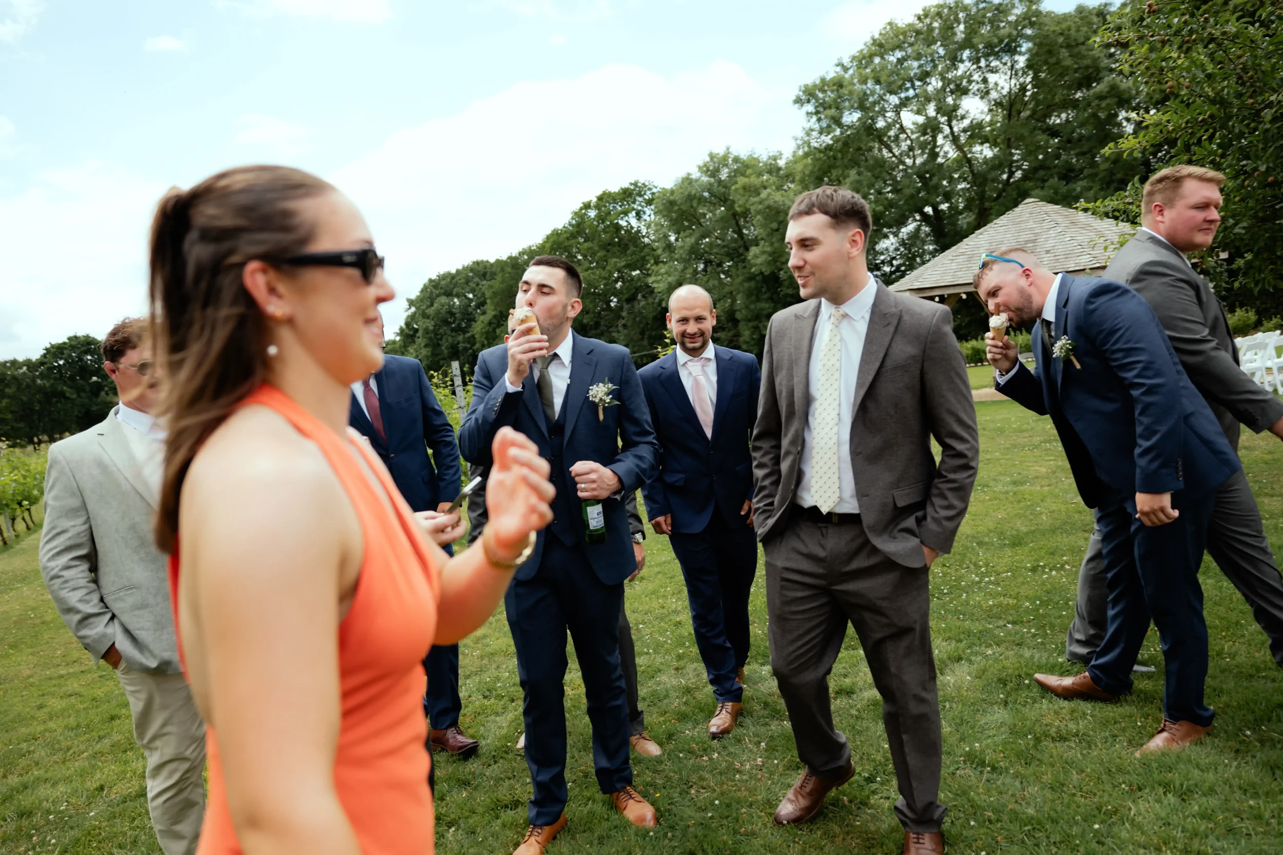 Group of people outdoors at a social gathering on a grassy area, with some holding drinks and eating, and trees and a gazebo in the background.