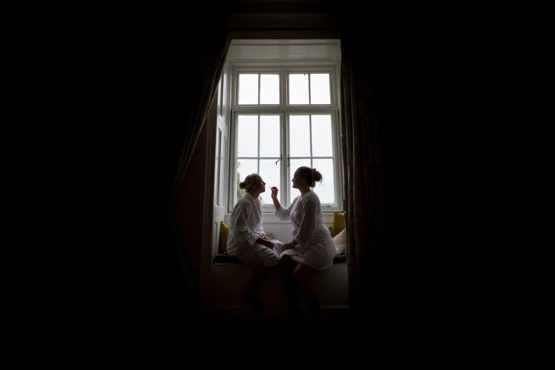 Two women in white robes sitting on a window seat, one applying makeup to the other, with a large window behind them and curtains on either side.