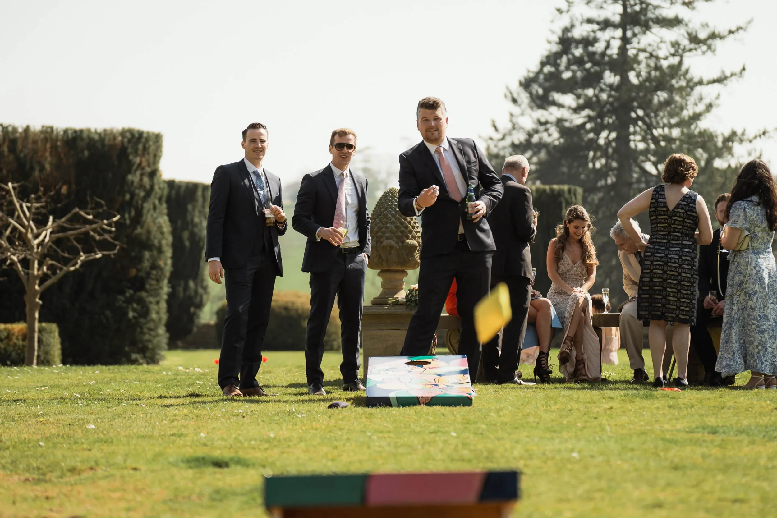 Group of people outdoors playing cornhole game on grassy lawn, with some dressed in formal attire, and others sitting and socializing in the background.