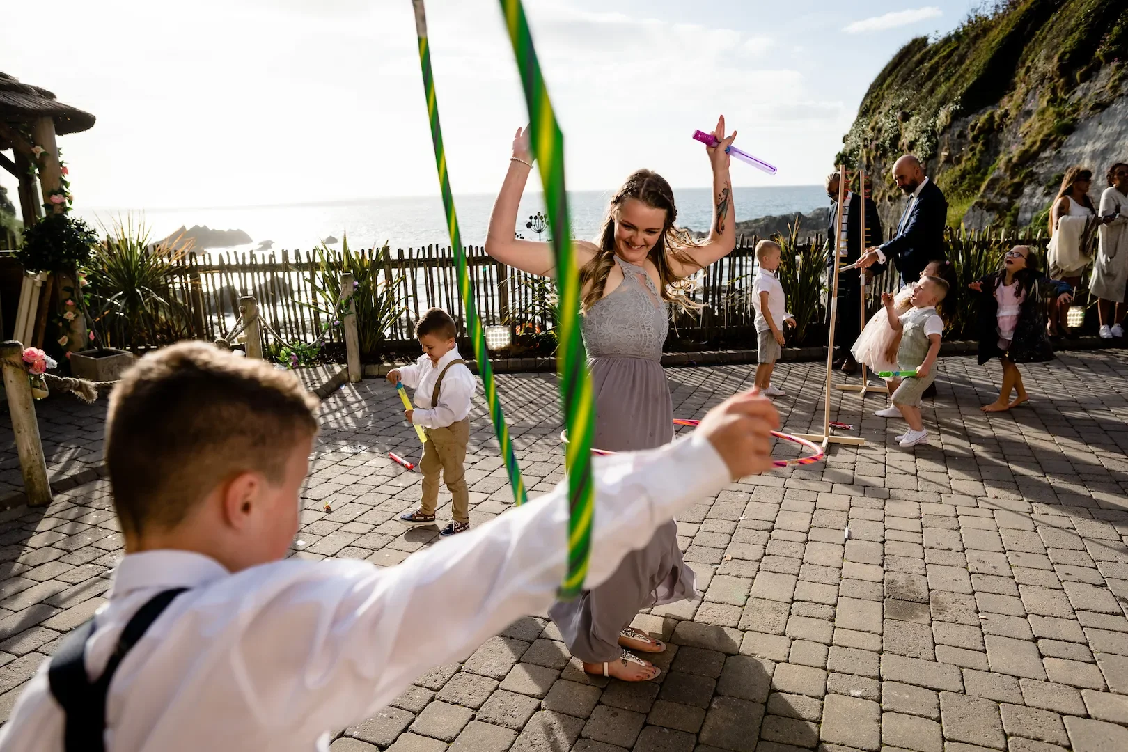 Children and adults at an outdoor celebration near the coast, with some playing and dancing, and a scenic ocean view in the background.