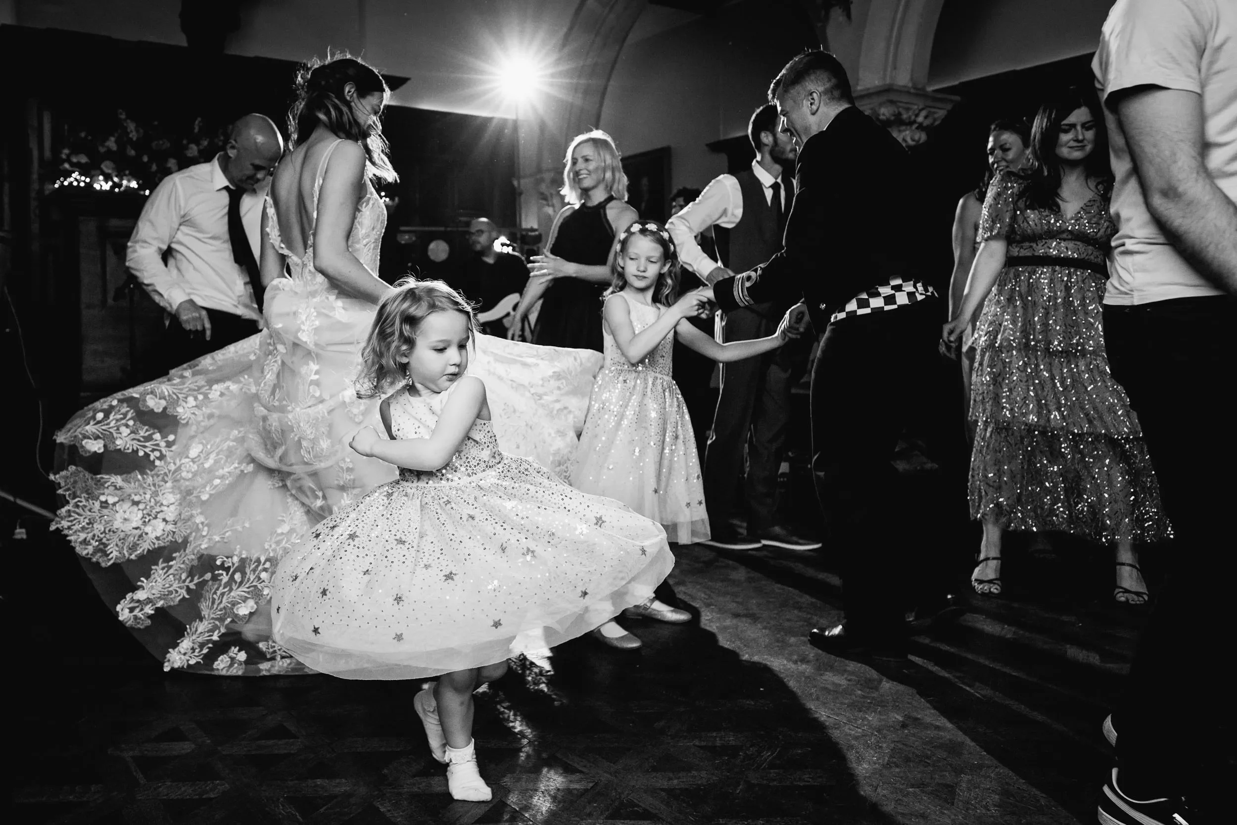 Children dancing at a celebration, with adults in the background, in a dimly lit room.