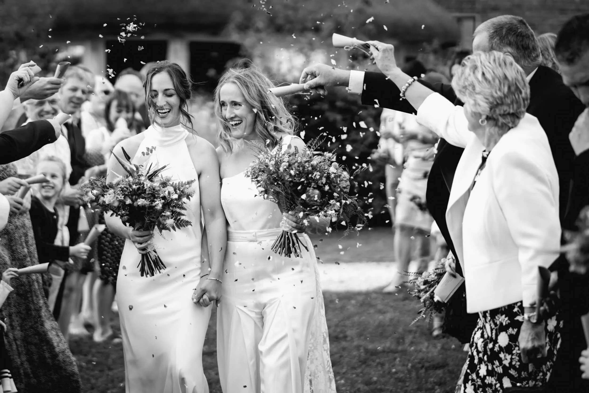 Two women in wedding dresses holding bouquets, walking through a celebration where people are throwing confetti at them. The scene is joyful and festive, with guests smiling and celebrating.