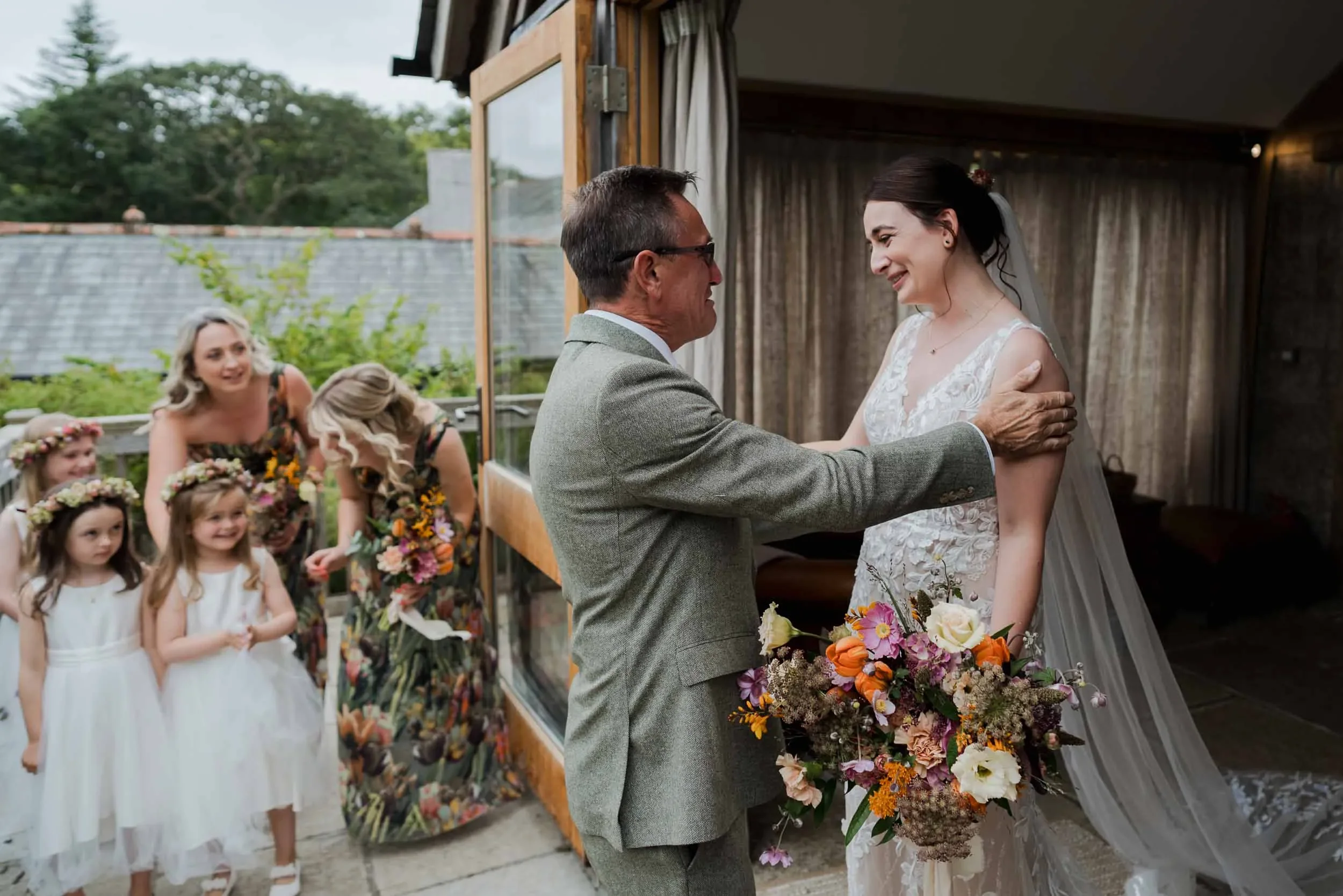 A bride smiling at her father during a wedding ceremony, with flower girls and bridesmaids in background.