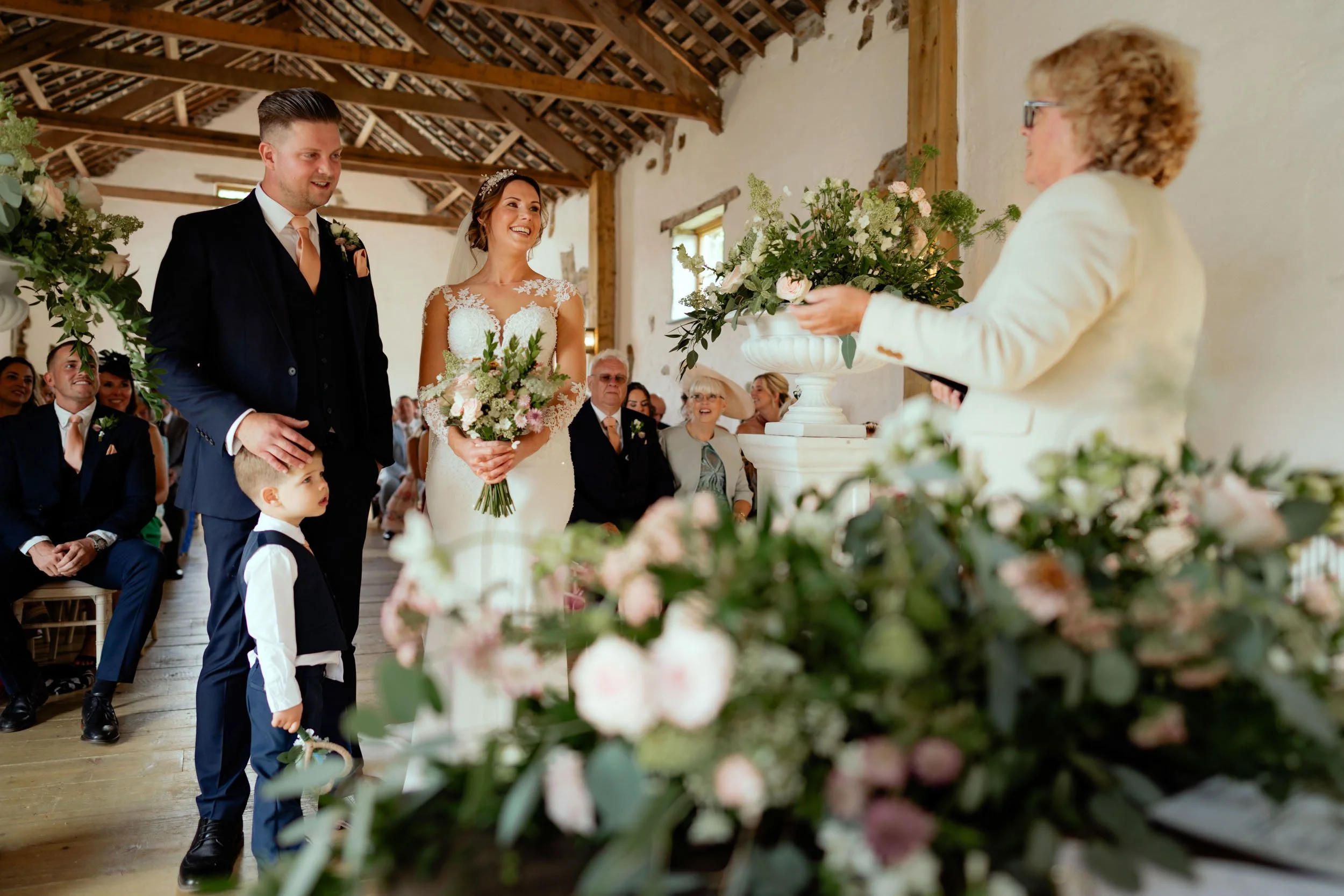 Bride and groom exchanging vows at their wedding ceremony in a rustic venue with floral decorations, while guests watch and smile.