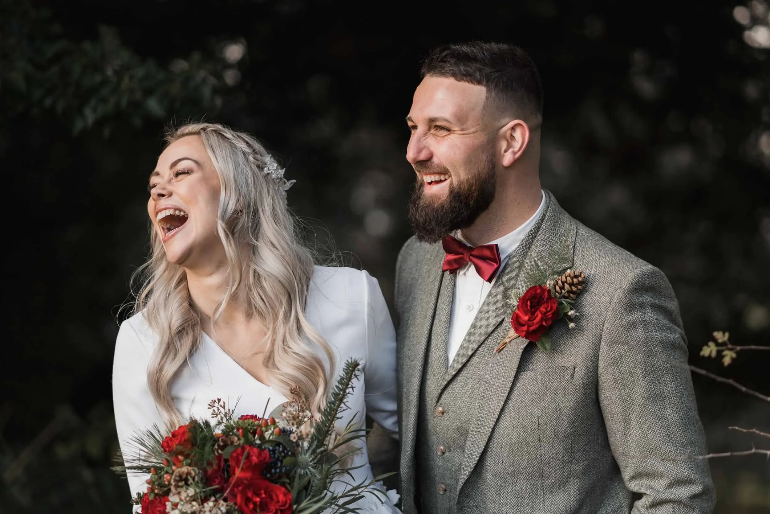 A bride and groom laughing and smiling outdoors, with the bride holding a bouquet of flowers and the groom wearing a gray suit with a red boutonniere and matching bow tie.